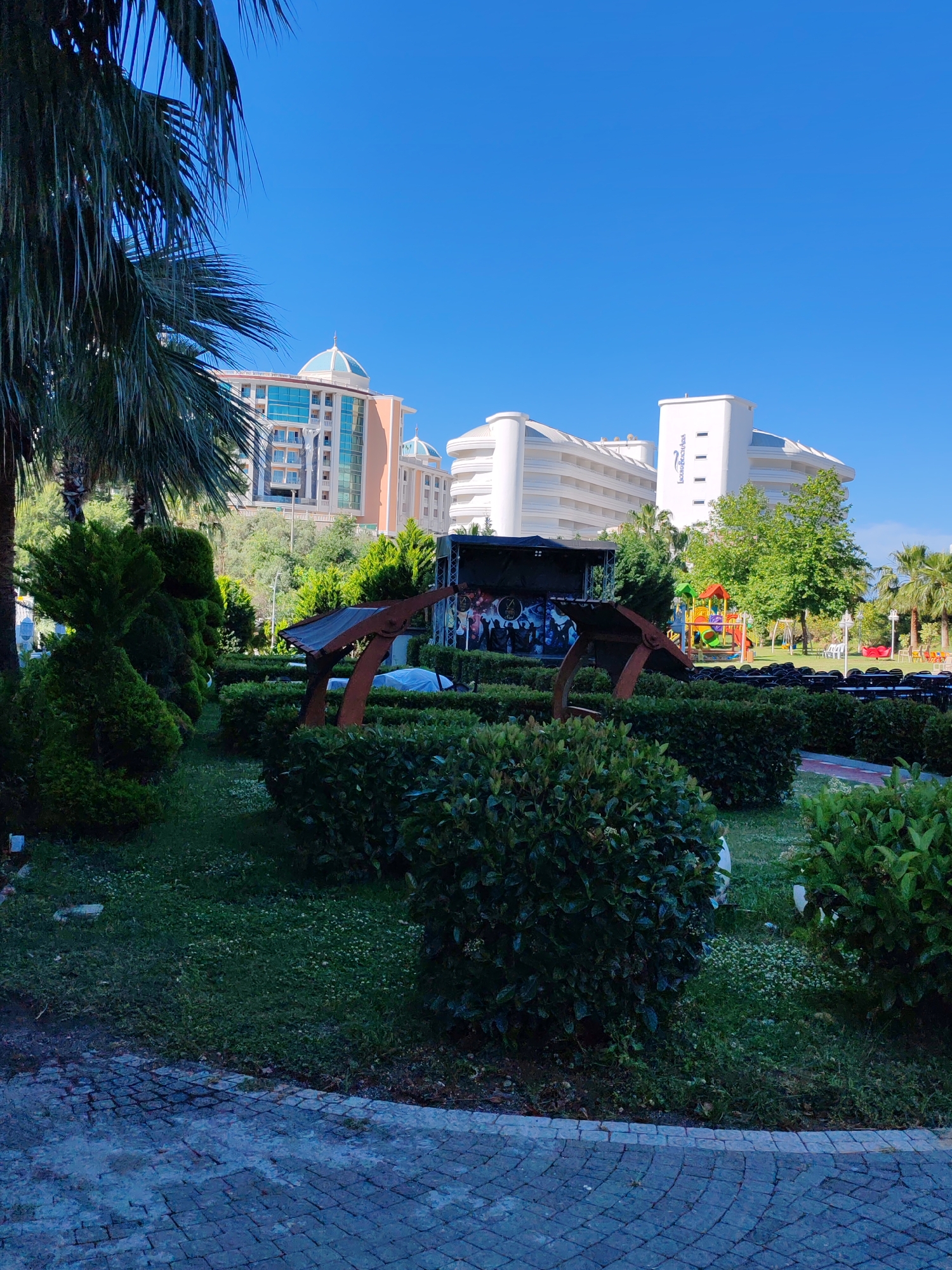 The image shows a park-like area in Alanya, Turkey, on a sunny day. In the foreground, there is a neatly trimmed hedge with a metal anchor-shaped structure. Beyond the hedge, there is a stage set up with rows of chairs facing it, suggesting an outdoor performance area. Further back, a playground with slides and swings is visible.
In the background, there are two large hotel buildings: one is a modern white building with curved architecture and a blue logo (possibly "Long Beach"), and the other is a taller, more ornate building with a light orange facade and a blue dome on top. Palm trees are scattered throughout the scene, contributing to the tropical atmosphere of the location. The area is paved with grey stone blocks in the immediate foreground. The scene is well-maintained and likely part of a resort or tourist attraction.