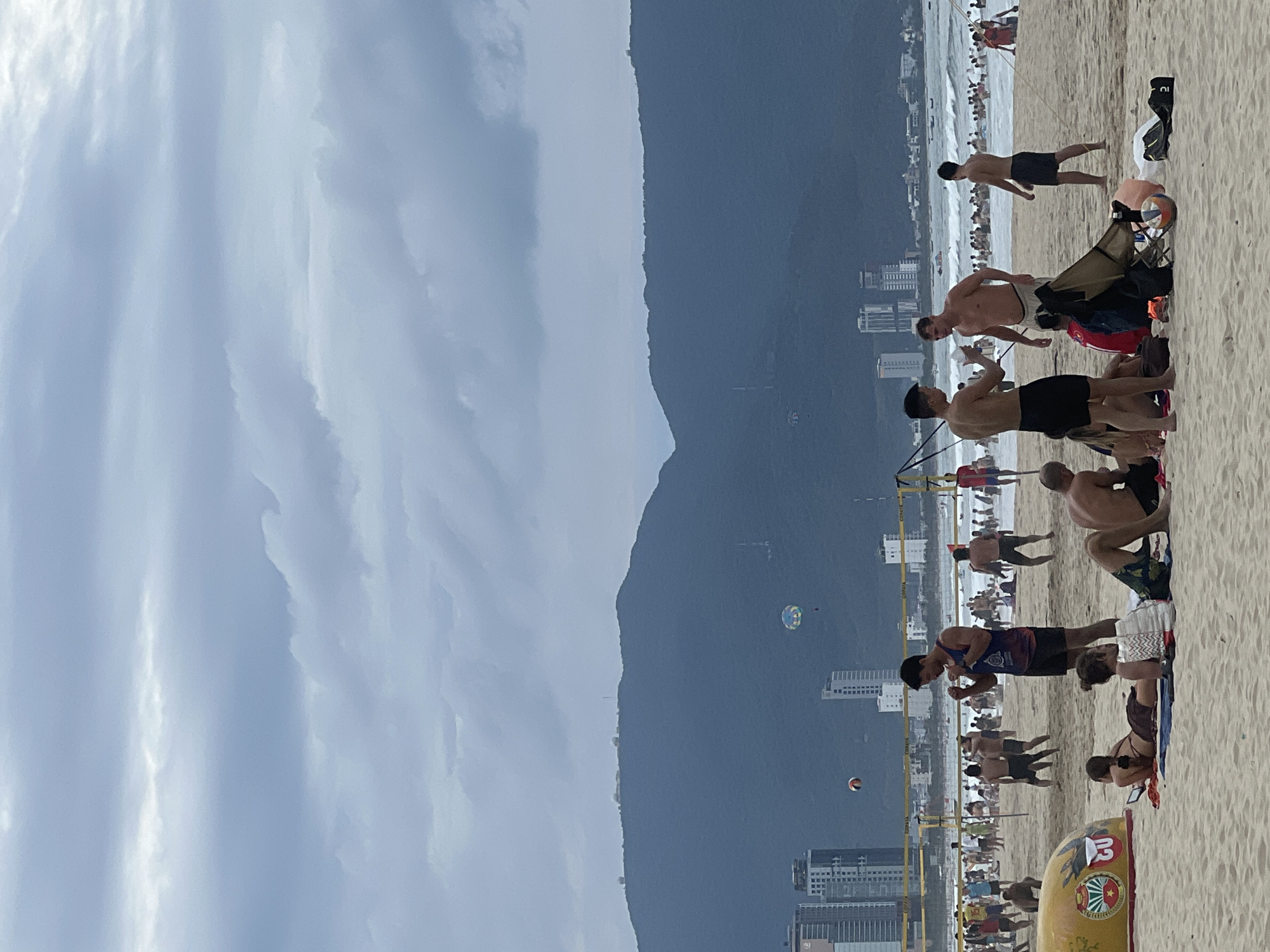 This image captures a bustling beach scene on a cloudy day, likely in Đà Nẵng, Vietnam, given the coastal landscape and architecture. In the foreground, several people are relaxing on the sand, some lying down and others sitting. A distinctive yellow and red boat-like object with emblems is visible on the left. Further back, a group of men are engaged in conversation or activity. A volleyball net stretches across the sand, indicating recreational use. Beyond the beach, a crowded ocean with numerous swimmers can be seen, with waves rolling in. In the middle ground, a city skyline with tall buildings rises, and in the background, a large, verdant mountain range dominates the horizon, with a paraglider visible in the sky above it. The overall atmosphere is one of leisure and outdoor activity.