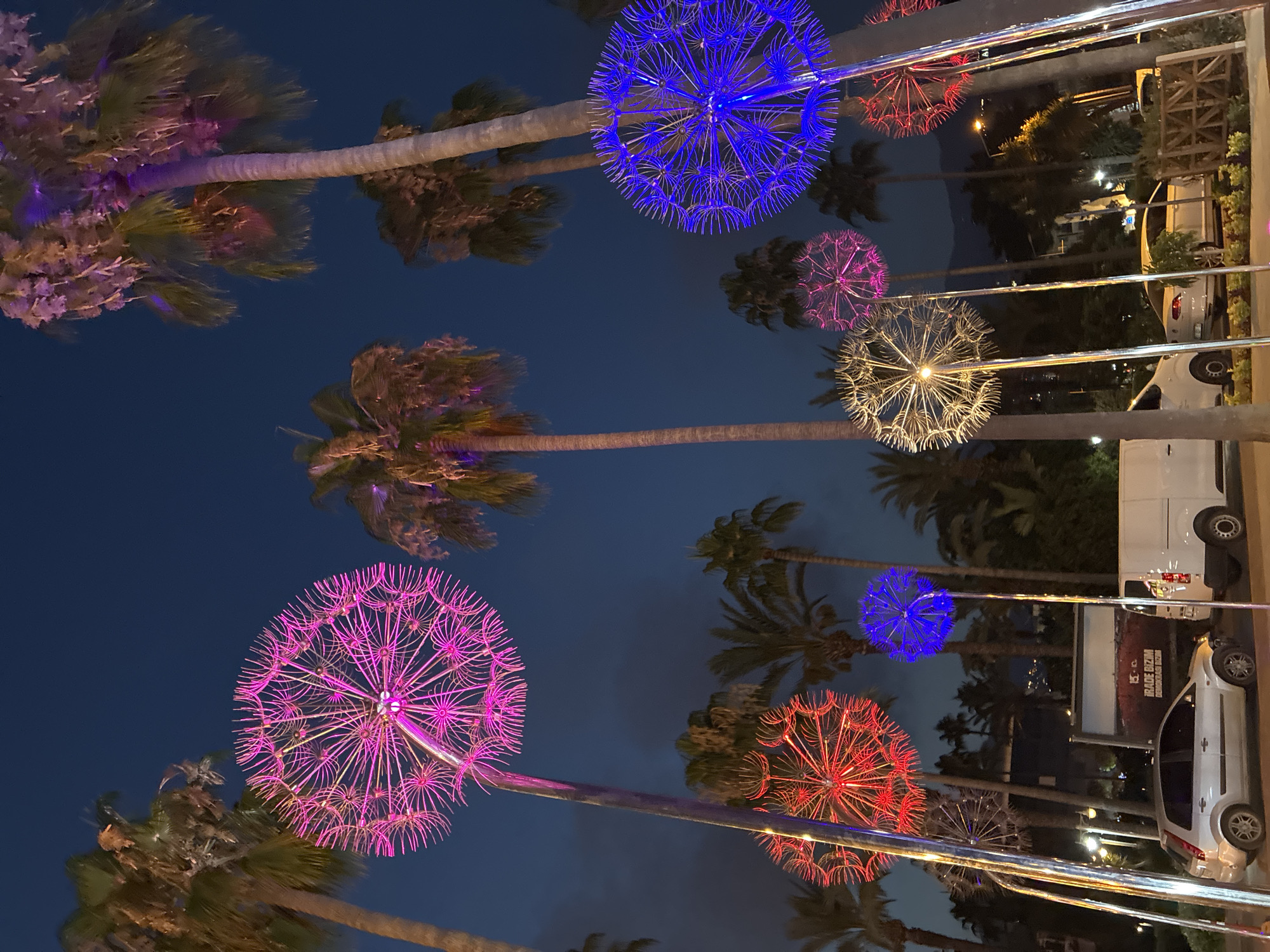In Alanya, Turkey, at dusk, a street scene features large, illuminated dandelion-like light sculptures. Palm trees with vibrant pink and purple lighting dot the background, contrasting with the deep blue twilight sky. A silver car is parked on the left, next to a white van. In the background, a billboard displays the Turkish words "BİZİM GELECEĞİMİZ" which translates to "Our Future". The lighting casts a colorful glow on the tropical foliage and the urban setting.