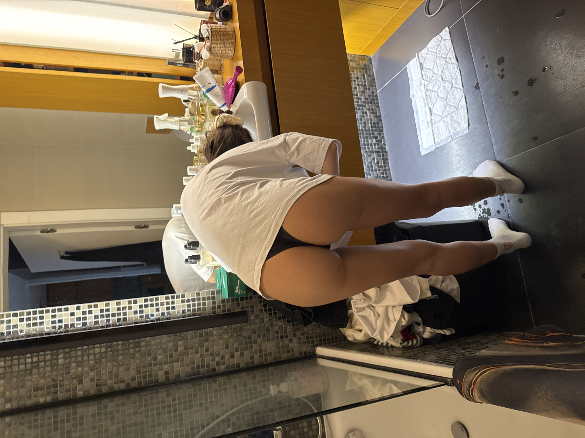The image depicts an indoor bathroom scene, primarily focused on one individual from a rear perspective. The person, appearing female, is bent over a white ceramic sink, with her head and hands largely out of view below the sink's rim. She is wearing an oversized white t-shirt that rides up, revealing black thong underwear. Her hair, dark in color, is tied back with a light-colored scrunchie. She wears white low-cut socks, with faint red lettering visible on the left sock.
The bathroom features walls clad in a mosaic pattern of small, square, metallic-toned tiles (greens, browns, grays, whites). The floor is composed of large, dark gray or black tiles, and several water droplets are visible on the floor near the person's feet. A white absorbent pad or mat is also on the floor.
To the left, a glass-paneled shower enclosure is visible, containing a white tub base and a clear/pink pump bottle. A pile of white and dark clothing or towels rests on the floor next to the shower enclosure.
The sink is part of a light brown wooden vanity or cabinet. The countertop surrounding the sink is cluttered with numerous toiletries, including various pump bottles, spray bottles, jars, tubes (one blue and white), a small woven basket, a pink hairbrush, and a dark cylindrical object (possibly a candle or diffuser).
A large mirror occupies the background, reflecting parts of the room. Within this mirror's reflection, another individual, or possibly a complex reflection of the primary subject, is visible, also wearing a white top and appearing bent over, possibly at a different sink or counter. The reflection also shows a white door partly ajar, revealing a darker room beyond, and vertical yellow/gold paneling or a frame on the far right.
