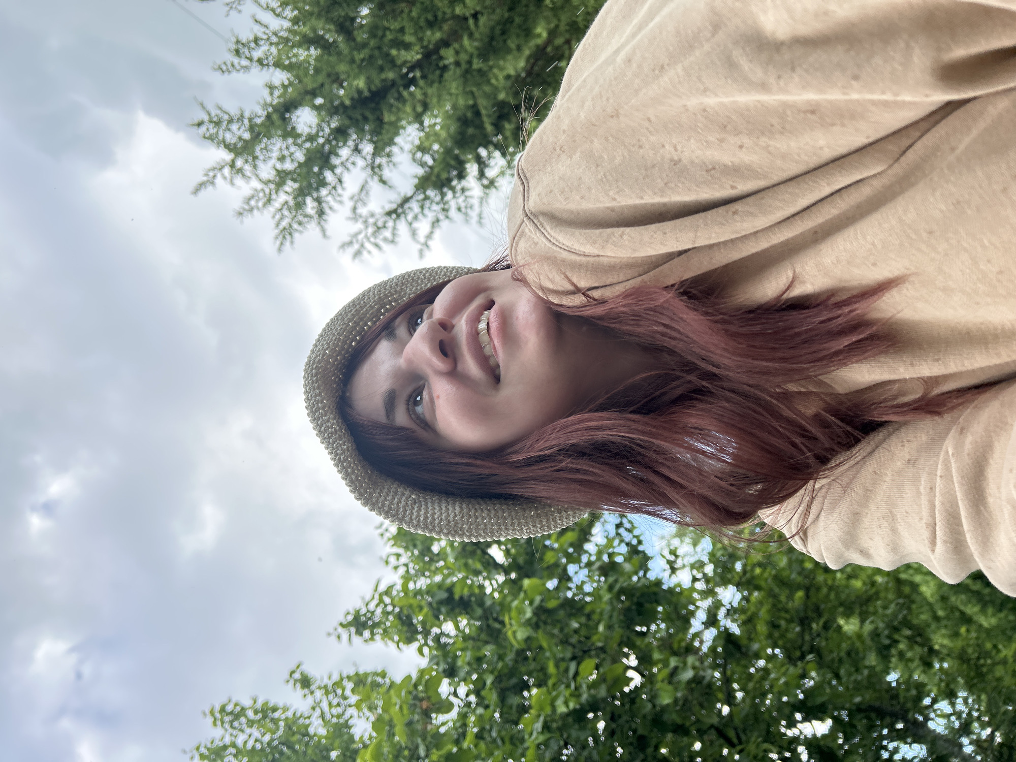The image is a rotated portrait of a young woman taken from a low-angle perspective, looking upwards. She is positioned on her side within the frame, smiling or smirking pleasantly. She has reddish-brown hair and is wearing a light-colored, possibly beige or straw, hat and a matching beige hooded sweatshirt. Her gaze is directed upwards and slightly to her right (relative to her body).
The background consists of lush green trees with varied foliage, some in sharp focus and others softly blurred, suggesting an outdoor natural environment. Above the trees, a cloudy sky with shades of gray and white is visible, indicating an overcast or partly cloudy daytime setting. The overall atmosphere appears relaxed and casual. There is no visible text. The location of this scene is Foki, Russia.