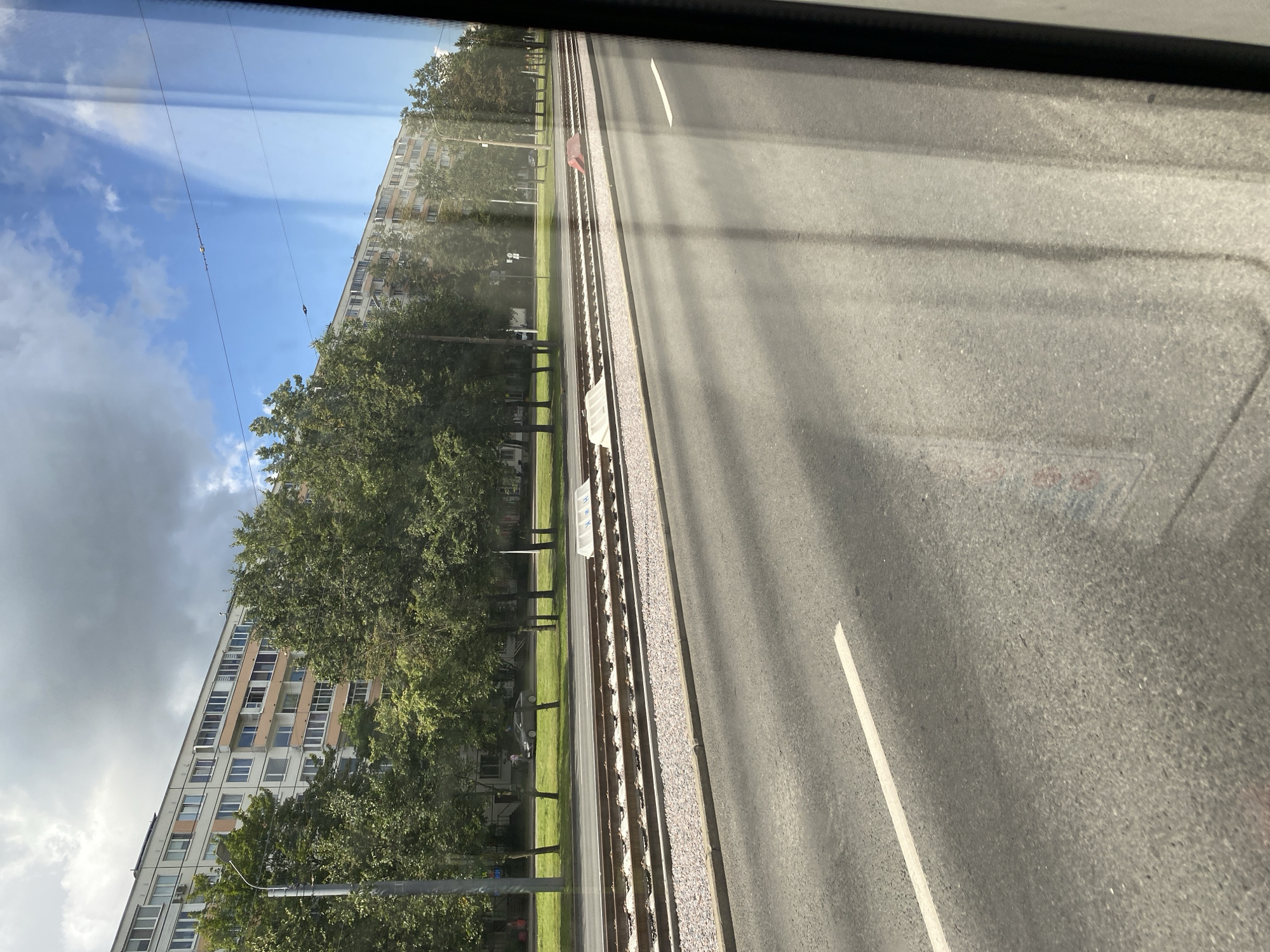 This image, taken from a window, shows a view of a street in Saint Petersburg, Russia. In the background, a long, multi-story apartment building with a light-colored facade and many windows runs horizontally across the frame. Trees with green foliage are situated in front of the building, casting shadows. In the middle ground, there are tram tracks with gravel ballast. A road with a white lane marking is visible in the foreground, with shadows from the tram wires falling across it. The sky is a mix of blue and white clouds. A silver car is parked on the side of the road, and a person is walking near it. Another person is seen on a sidewalk further down the street. A white barrier and a red barrier are present near the tram tracks.