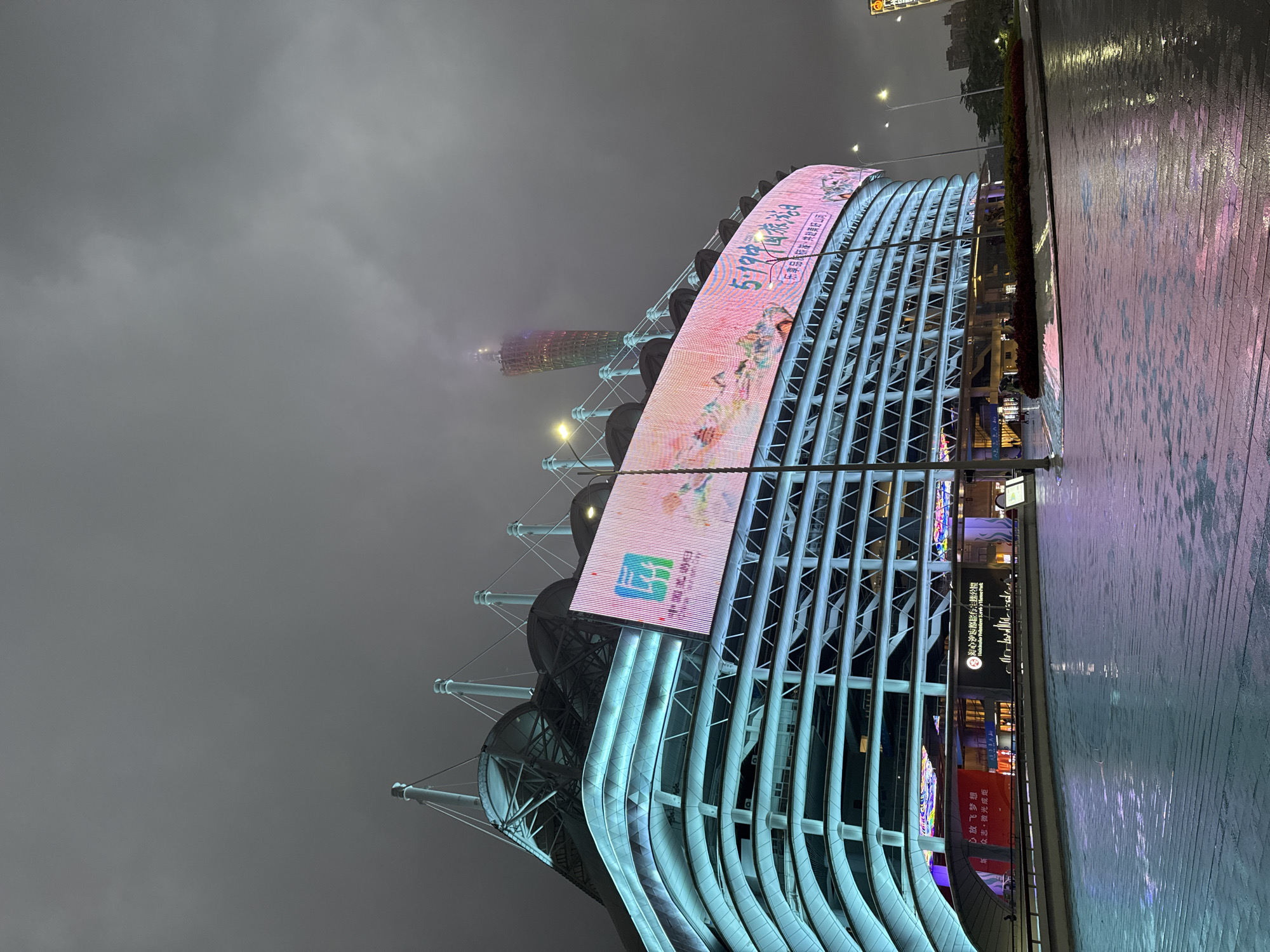 The image shows a modern building in the Tianhe District of Guangzhou, China at night. The building is characterized by a curved, illuminated screen displaying images and Chinese text, supported by a framework of steel beams and cables. The Canton Tower is partially visible in the background, obscured by heavy fog. The foreground features a wet, reflective sidewalk, indicating recent rain. The overall atmosphere is urban, with a focus on the contemporary architecture and the visual impact of the illuminated displays.