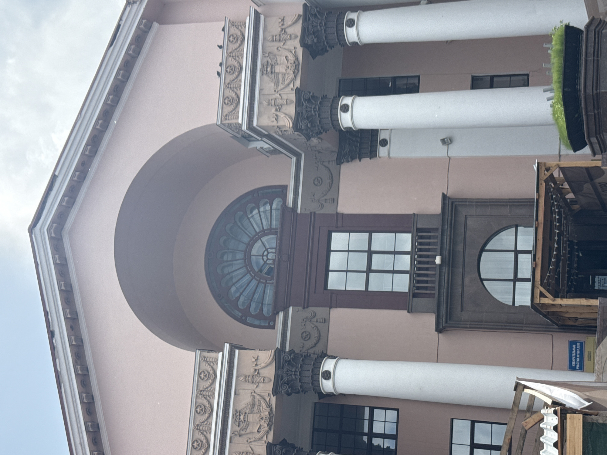 The image depicts the upper and mid-sections of the Yekaterinburg Opera and Ballet Theatre, located at 46 Prospekt Lenina in Yekaterinburg, Russia. The building is characterized by its light-pink facade and grand Neoclassical/Stalinist Empire architectural style.
In the upper center, a large, recessed archway frames an ornate semicircular window with a radiating fan-like spoke design. Below this, a large rectangular window features a dark frame and multiple panes, surmounted by a small, dark balcony with horizontal railings.
Flanking the central archway are two prominent sections, each supported by two robust, white fluted columns with dark, intricately carved Corinthian or Composite capitals. These columns support ornate, light-brown friezes. The frieze on the left features relief carvings including a lighthouse, a ship, a torch, and the year "1917." The frieze on the right displays decorative stars and wreaths. Several dark birds, likely pigeons, are perched on the top edge of the right frieze.
Below the left frieze and columns, multiple dark-framed rectangular windows with grid patterns are visible. On the lower left, a blue rectangular sign is affixed to the wall, bearing white text that reads "ИЗБИРАТЕЛЬНЫЕ УЧАСТКИ №1267, 1268" (Electoral Precincts No. 1267, 1268) and "РОСПРОМСТРОЙ" below. This area is partially obscured by temporary wooden scaffolding and white tarpaulin.
On the lower right, a large black planter filled with green grass and several small metallic posts rests on a dark stone pedestal. Additional temporary wooden structures are present in the lower right foreground, obscuring the base of the building. The sky above is overcast. No individuals are visible in the scene.