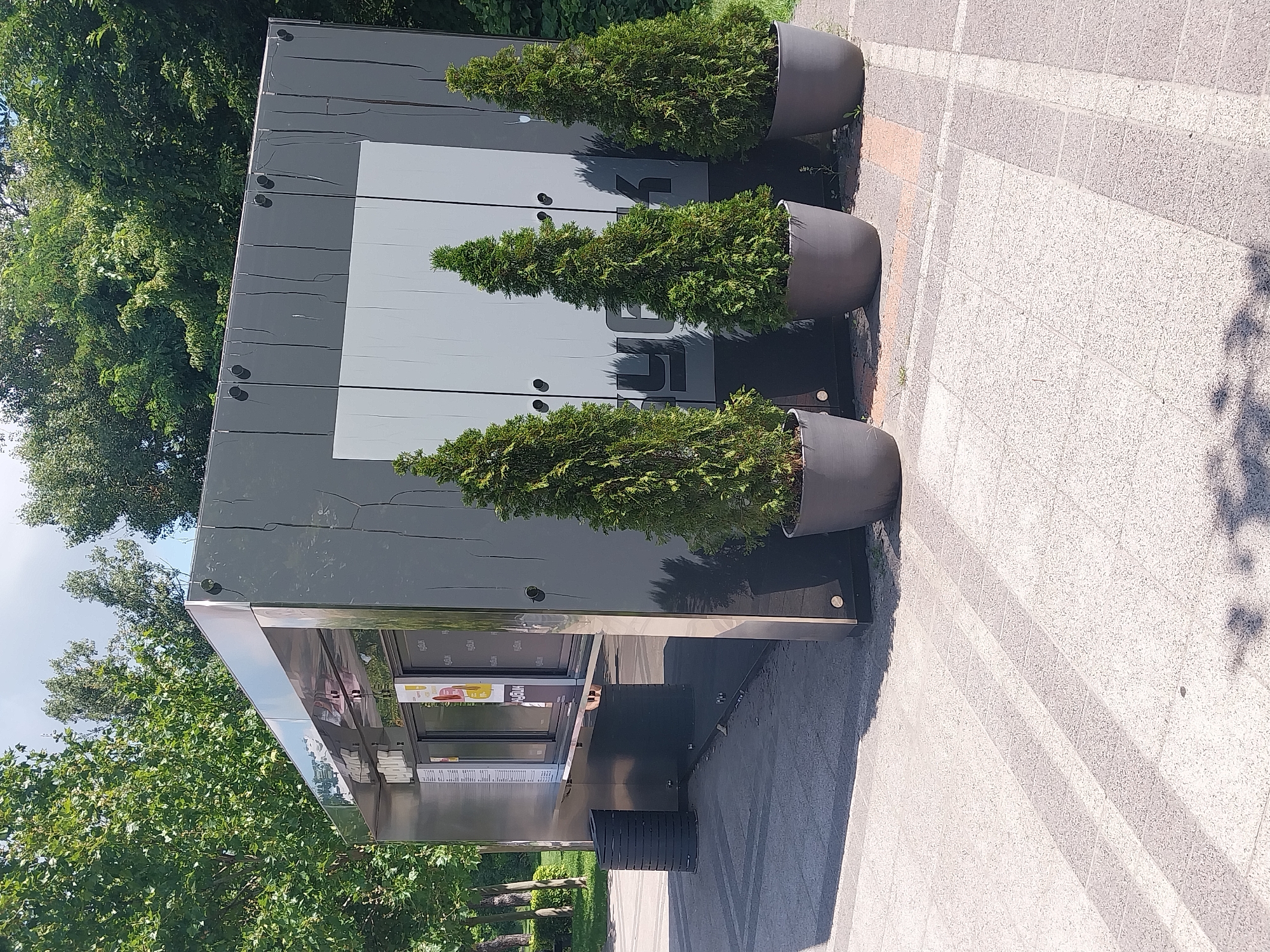 In Dnipro, Ukraine, a modern, gray food kiosk with a reflective silver trim is situated on a paved outdoor area. Three conical evergreen trees in dark gray planters stand in front of the kiosk. The kiosk features a service window and a sign with Cyrillic lettering. To the left of the kiosk, a black cylindrical trash receptacle is visible. The surrounding environment includes green trees and a bright sky. The pavement is composed of interlocking stone tiles with visible shadows from the trees and structure, indicating a daytime scene.
