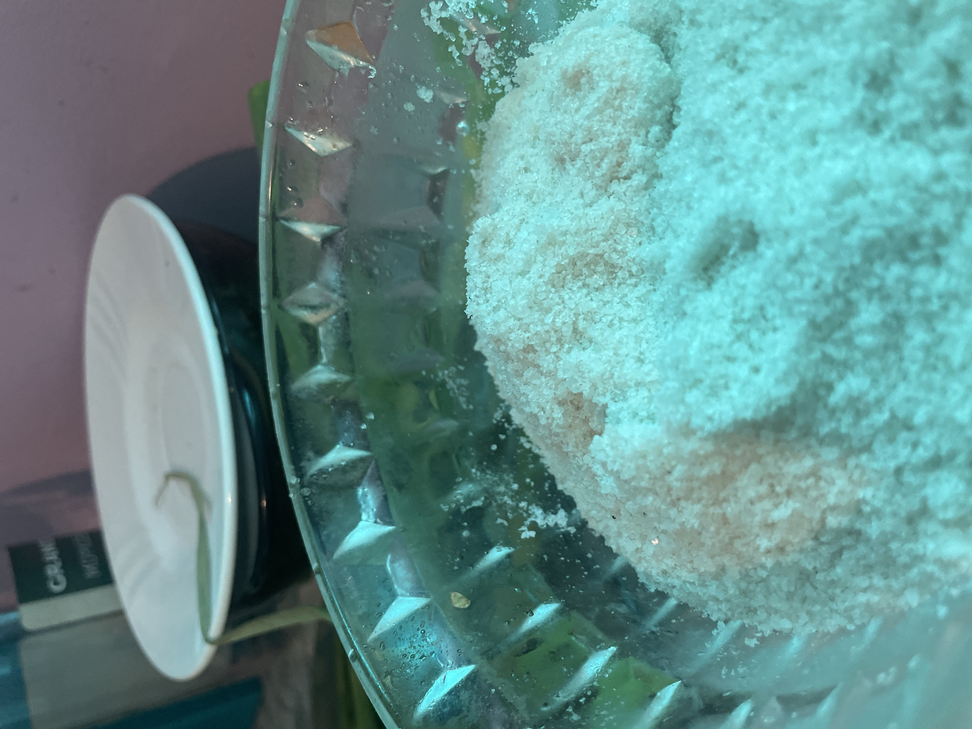 A close-up, slightly blurry, high-angle shot reveals a clear glass bowl containing a large, compacted mass of white granular material, possibly salt or sugar. The bowl features a textured exterior with diamond-shaped indentations. To the left, partially obscured, are a white plate stacked on a black object, potentially a container or another plate. The background is indeterminate, showing hints of green and pink. It is impossible to determine if this image was taken in Olginka, Russia.