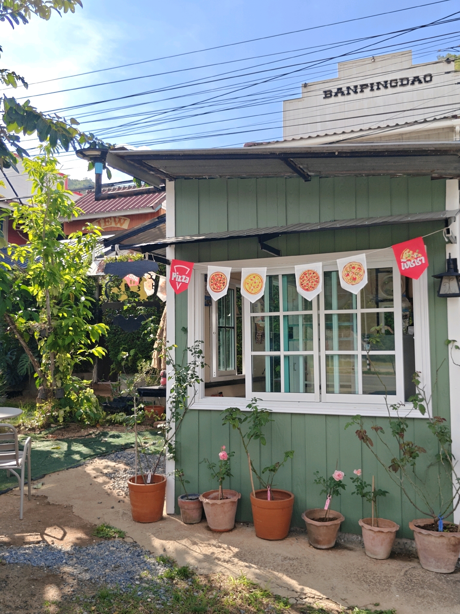 The image depicts the exterior of a small, green, wooden building situated in Khao Yai, Thailand. The building has a corrugated metal roof and a large, white-framed window with multiple panes. A string of small pennant flags hangs above the window; some are red and white with pizza graphics, while one is red with Thai writing and a pizza graphic. To the left of the building, a tree with green leaves is visible. In front of the building, several potted plants, including rose bushes, are arranged on a concrete patio. The ground in front of the patio is a mixture of gravel and sand. In the background, other buildings with red roofs and a sign with the text "BANPINGDAO" are visible. The sky is clear and blue. No people are present in the image.