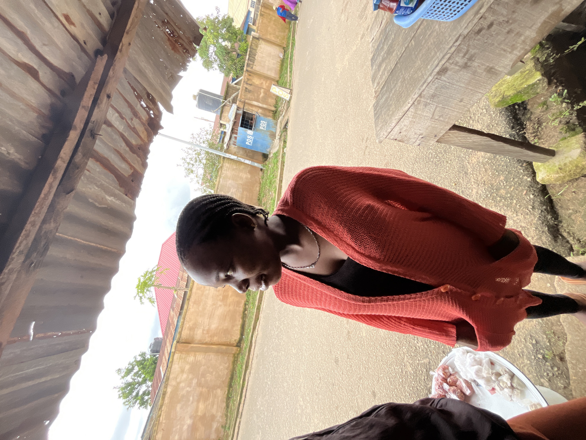 A medium shot captures a young Black woman with braided hair, wearing a coral-colored knitted cardigan over a black top. She stands to the right of the frame, her body angled towards the left, and her gaze directed off-camera to the left. Her hands are placed in the pockets of her cardigan. To her left, another person, partially visible, wears a dark brown top and orange pants.
In the foreground, a white tray holds wrapped food items. To the right, a weathered wooden table is set up. The background shows a street with a light brown wall bordering it. Beyond the wall, there are buildings with red roofs, trees, and a blue structure with "POS" written on it. A water tank is visible on top of a building. Further down the street, two figures, one in a blue and white outfit and another in a pink outfit, are walking away from the camera. The scene appears to be an outdoor market or roadside stall in the city of Jos, Nigeria, under an overcast sky. The corrugated metal roof of an overhead shelter is visible at the top of the frame.