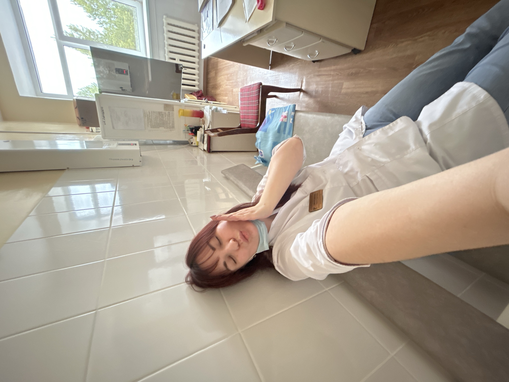 A young woman with reddish-brown hair, wearing a white lab coat over blue trousers and a light blue surgical mask pulled down to her chin, is lying on a white tiled floor. She appears to be resting or playfully posing due to exhaustion, with her eyes closed and one hand covering her forehead. She is leaning against a low, grey bench or step.
The setting is an indoor room, likely an office, clinic, or laboratory, possibly in Фоки, Russia. Bright natural light streams in through a large window, revealing green foliage outside, indicating it is daytime. A white radiator is visible below the window. In the background, there's a white desk with drawers, a wooden chair with a red and white striped cushion, and various items including boxes, a blue bag, and shelves. Two white rectangular devices, possibly medical or laboratory equipment, are placed near the window. One has a small red light illuminated on its screen. Text is partially visible on one device, possibly "DIGITALLY," and on another, "THERMO SWIFT" might be discernible. The woman wears a small, unreadable name tag on her lab coat. The overall scene conveys a moment of casual rest in a professional environment.