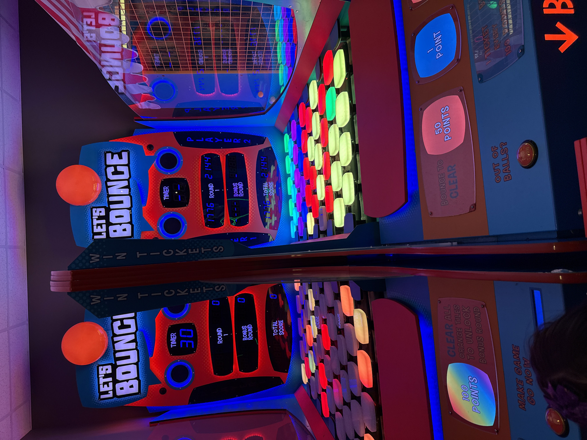 The image shows a "Let's Bounce" arcade game, potentially at an entertainment center in Columbus, Ohio, USA. The game is designed for two players and involves bouncing balls to hit lit-up tiles. The display shows a timer, round number, bonus round status, and total score for each player. Instructions on the machine explain the point system and how to earn bonus rounds. An orange ball is present on one side of the game.