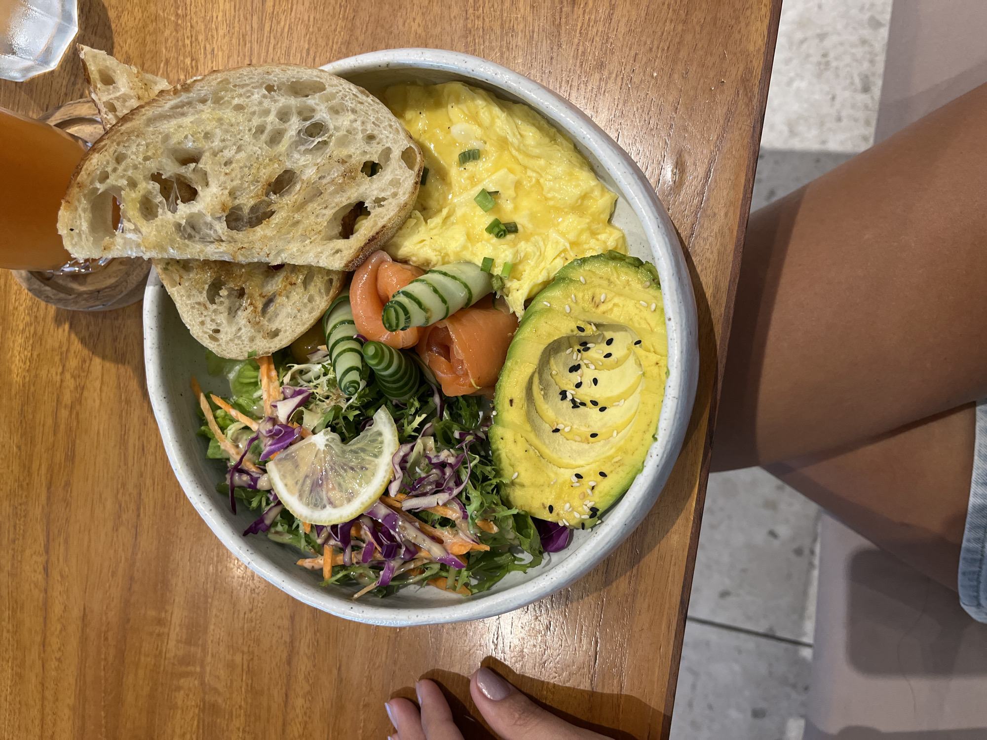 The image shows a breakfast bowl on a wooden table, likely in Kuta Selatan, Indonesia. The bowl contains scrambled eggs, sliced avocado with sesame seeds, smoked salmon, spiral-cut cucumbers, a slice of lemon, and a salad with shredded carrots and purple cabbage. Two slices of bread lean against the bowl. A glass of orange juice is visible in the background. A person's leg and hand are also in the frame, indicating they are sitting at the table.