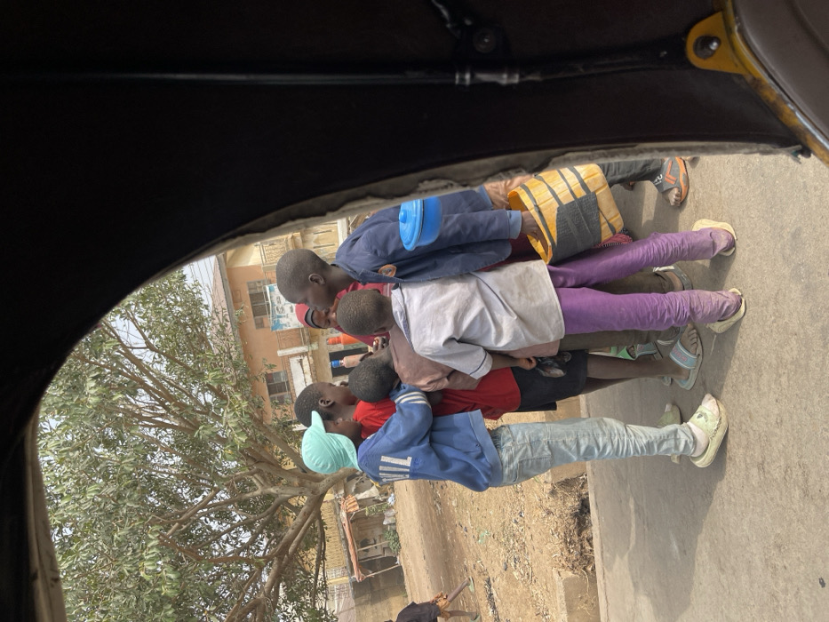 The image, framed from within a vehicle, depicts an outdoor scene in Samaru, Nigeria, during daylight hours. A group of seven children are clustered on a paved surface.
In the foreground, a child with their back mostly to the viewer wears a light grey shirt, purple trousers, and light-colored slip-on shoes. To their left, a child in a light blue cap, blue long-sleeved shirt, and light blue jeans stands, with one arm around another child wearing a red shirt. Partially obscured among the cluster is a female child wearing a reddish head covering. To the right of the central cluster, another child in a dark blue jacket holds a yellow plastic jerrycan or bucket with black strapping in their left hand, and a blue lidded bowl is hooked onto their left shoulder. This child also wears dark trousers and dark sandals. Other children are partially visible within the huddle, wearing various tops and shorts/trousers. The children appear to be interacting or observing something within their immediate group. Footwear varies from sandals to slip-on shoes.
To the left of the paved area is an unpaved dirt ground with some sparse vegetation. In the mid-ground, a large deciduous tree with mostly bare branches is prominent. In the background, a two-story building with a light brown/orange facade, featuring multiple windows and a balcony with a dark railing, is visible. Adjacent structures are partially seen. The sky is clear, suggesting fair weather. Another lone figure is partially visible on the dirt path in the far left background.