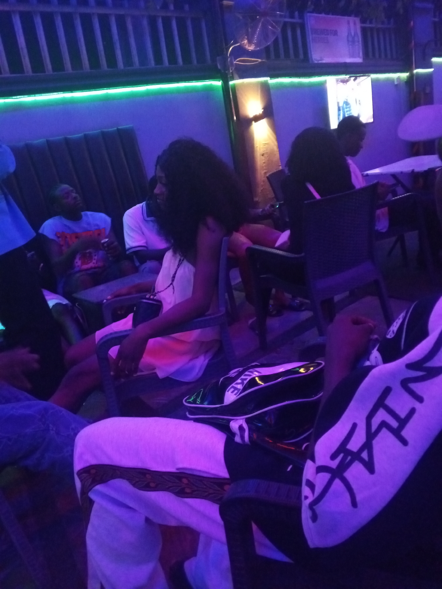 The image depicts a dimly lit outdoor venue in Lokoja, Nigeria. Several people are seated in chairs around tables. A woman with long dark hair wears a light-colored dress. Two men are seated at a table in the background. Green neon lighting is visible. A Nike duffel bag is partially visible in the foreground. The overall atmosphere appears to be that of a casual social gathering.