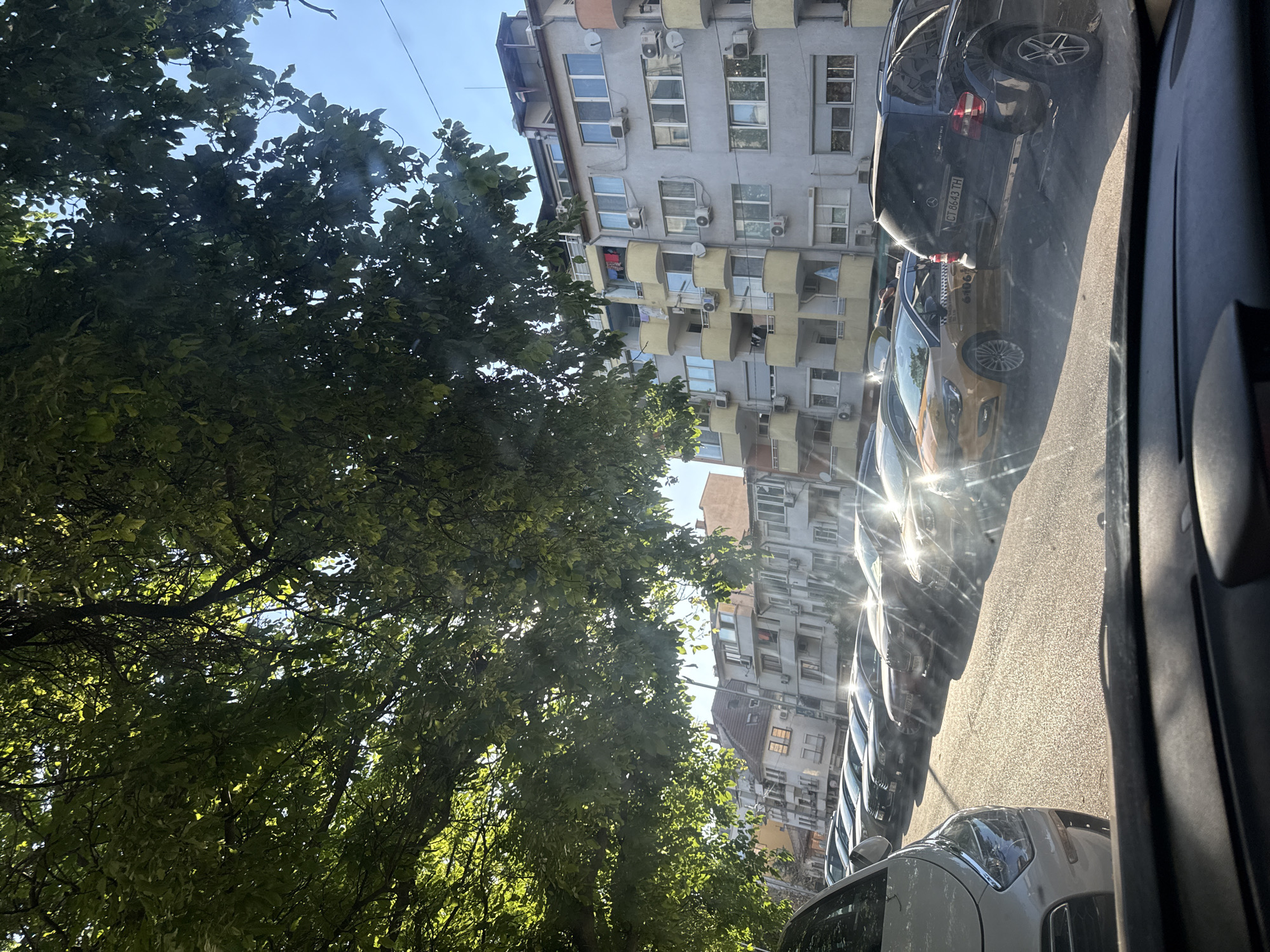The image depicts a street scene in Stara Zagora, Bulgaria, likely a residential area, on a sunny day. Lush green tree branches overhang the foreground, casting dappled sunlight onto a row of parked cars. The cars, mostly sedans and SUVs, are parked along the side of a paved road, with a bright yellow taxi with markings "CT 6106" visible among them. In the background, a multi-story apartment building dominates the scene. The building is light gray with several balconies, many of which have air conditioning units installed. Clothes are seen hanging on a clothesline on one of the balconies. The sky is clear and blue, indicating good weather. The overall impression is a typical urban environment in Eastern Europe, captured on a bright, sunlit afternoon.