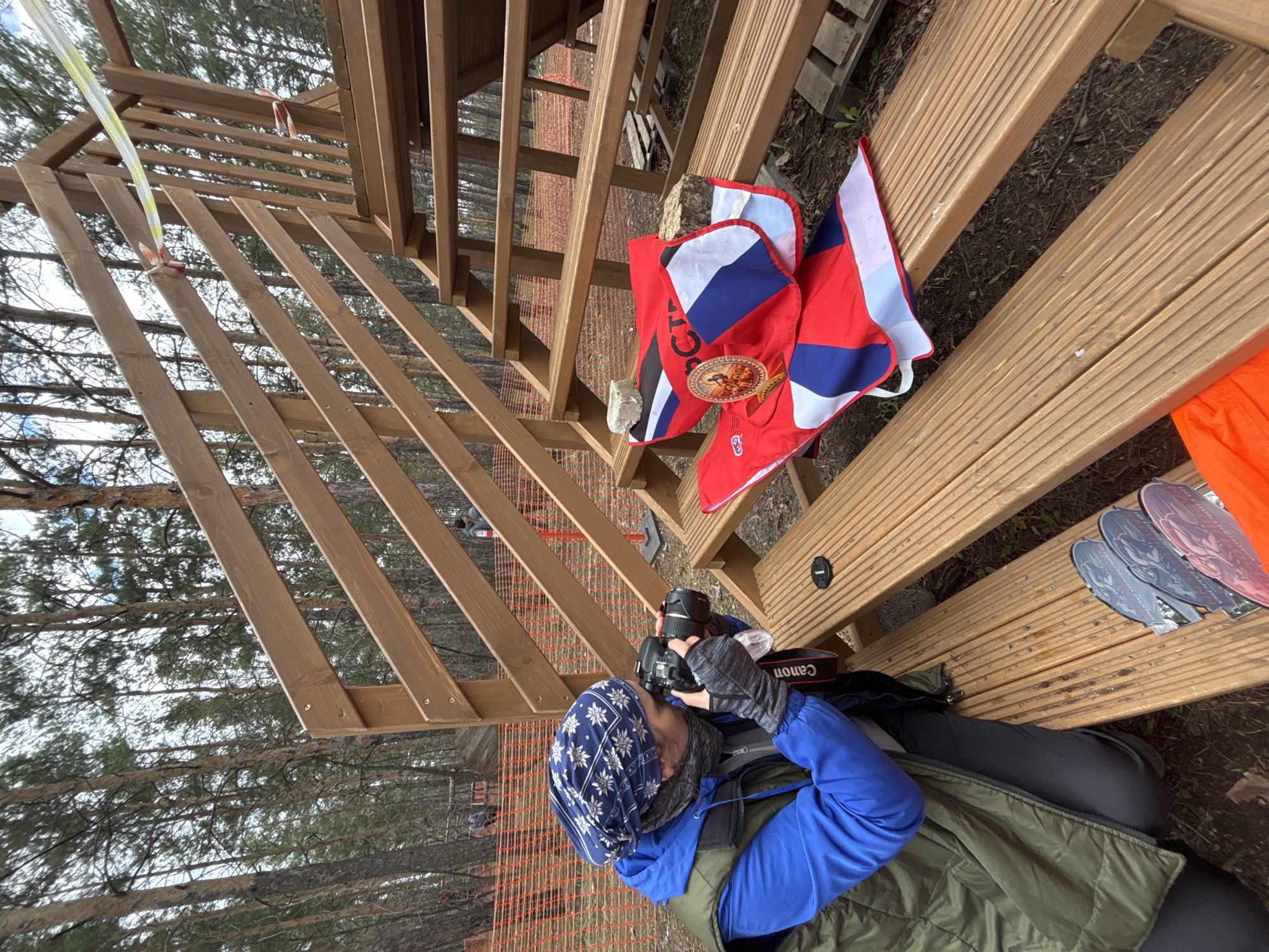 This image depicts an outdoor scene at an event in a forested area, located in Orenburg, Russia. The central subject is a person, likely a photographer, positioned on a robust wooden structure that resembles bleachers or a viewing stand. They are dressed in layered clothing including a blue jacket, a green or khaki vest, black pants, and a blue and white patterned head covering (bandana), along with dark gloves, suggesting cool weather. The person is actively engaged in photography, holding a black Canon DSLR camera equipped with a substantial lens, aimed towards an unseen subject.
To the right of the person, on the same wooden structure, several items are arranged. These include multiple small flags, predominantly red, white, and blue, consistent with the Russian national flag. One flag prominently displays partial text, "РОС..." (likely "РОССИЯ"), and a golden emblem featuring a double-headed eagle, a symbol associated with the Russian coat of arms. A black camera lens cap is also visible nearby. Further to the right, a bright orange piece of fabric and several elongated, grey, stylized objects, possibly medals or awards, are laid out. A wooden pallet can be seen in the background near these items.
The background scenery shows a dense, wooded environment, indicative of a pine forest, under a bright, possibly overcast daytime sky. An orange mesh safety fence or barrier is discernible through the gaps in the wooden structure, suggesting a demarcated area for an event such as a race, competition, or public gathering. The overall setting implies an organized outdoor event, with the photographer positioned to capture the unfolding activities. The visible text includes "Canon" on the camera strap and "РОС..." on one of the flags.