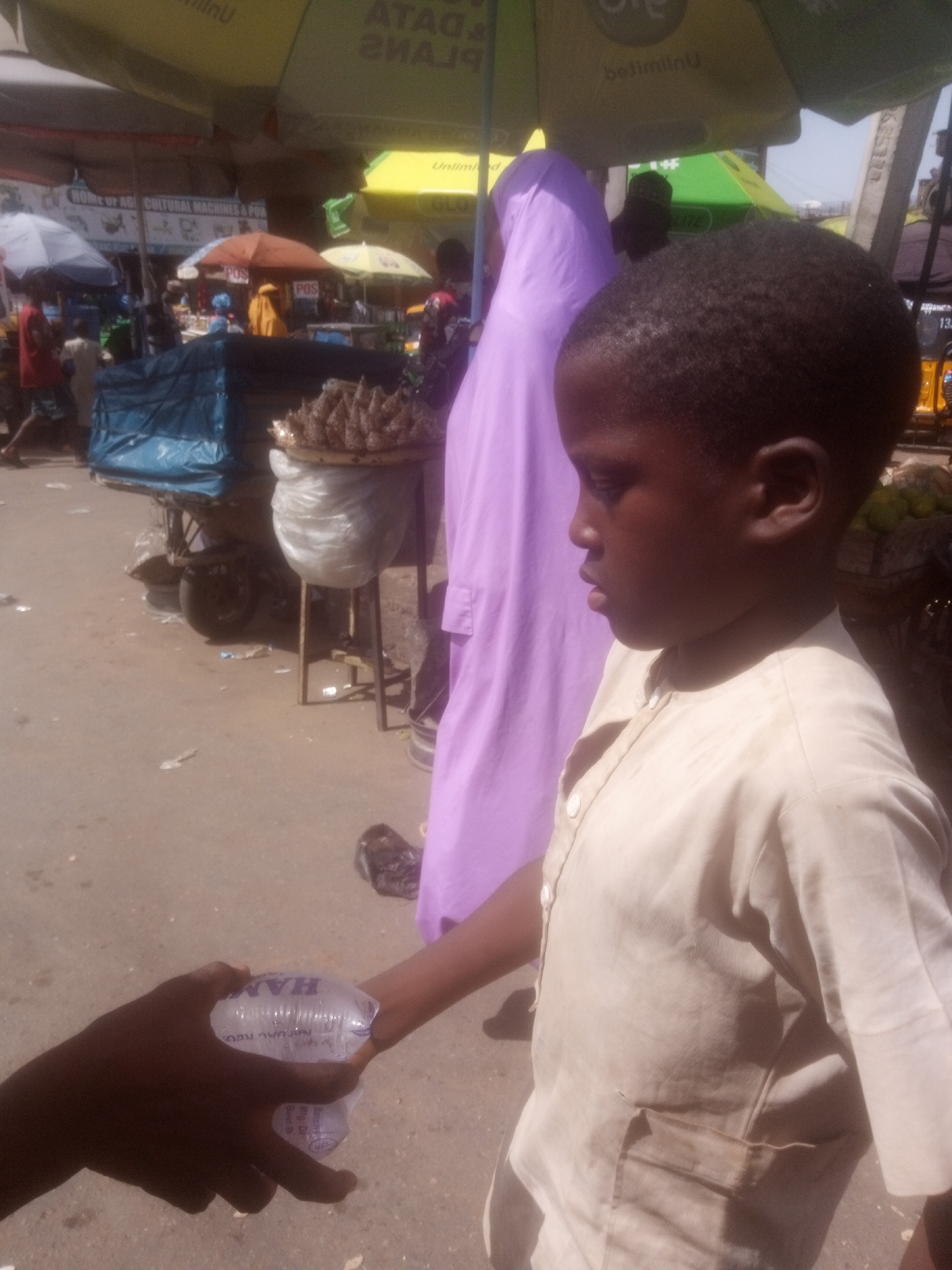 This image captures a bustling market scene in Maiburiji, Nigeria, bathed in bright sunlight, suggesting it's daytime. The foreground is dominated by the profile of a young boy wearing a light-colored, collared shirt. His gaze is directed downward, out of frame. In his hand, he holds a small, clear plastic sachet of what appears to be water, with visible text on it. Another hand, darker in skin tone, is reaching towards the sachet, possibly to take it or complete a transaction.
Behind the boy, a woman in a long, flowing lavender garment is partially visible, adding to the sense of a crowded public space. The background reveals a vibrant market atmosphere with various stalls and structures. Several colorful umbrellas, one yellow and one orange, provide shade over the vendors. In the mid-ground, a cart is laden with goods covered by a blue tarp, and a display of small, dried fish is arranged on a stand.
The ground is a dusty, uneven surface, typical of an outdoor market. Litter is visible on the ground, indicating the activity of the marketplace. Signs with text like "HOME OF AG! CULTURAL MACHINES & POR", "Unlimi", and "POS" can be seen, providing clues about the commercial nature of the location. The overall impression is one of everyday commerce and daily life unfolding in a Nigerian market.