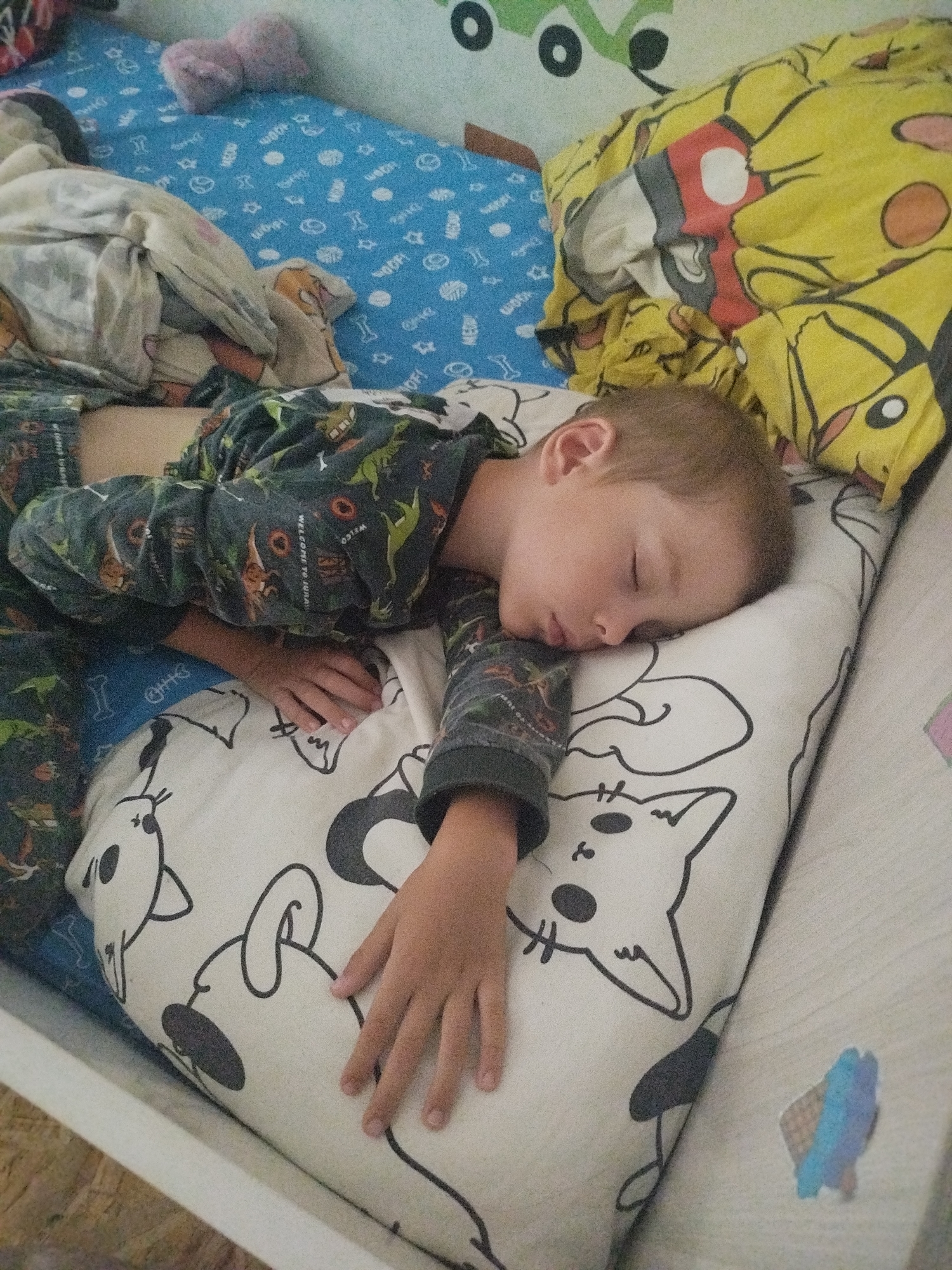 A young boy sleeps on his side in a bed. He is wearing pajamas with a dinosaur print. The bed has a blue sheet with white cartoon patterns, a yellow pillow with a Pikachu print, and a white pillow with black cat drawings. A light pink plush toy is visible on the bed. The scene appears to be indoors, likely a bedroom. The location is in the city of Zavodoukovsk, Russia.