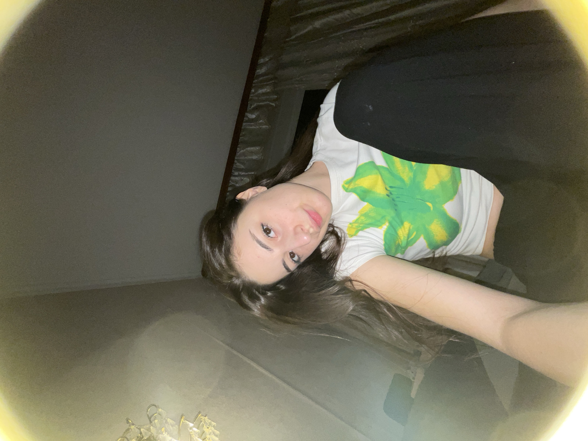 A young woman is visible in the center of the frame, wearing a white t-shirt with a large green and yellow floral design, and black pants. She is positioned at an angle, looking directly at the camera. Her hair is dark and falls loosely around her face and shoulders. The environment appears to be indoors, with a plain wall in the background and a textured ceiling visible above. Given the lack of specific architectural details or contextual clues, it is impossible to definitively pinpoint the location as Moscow, Russia.