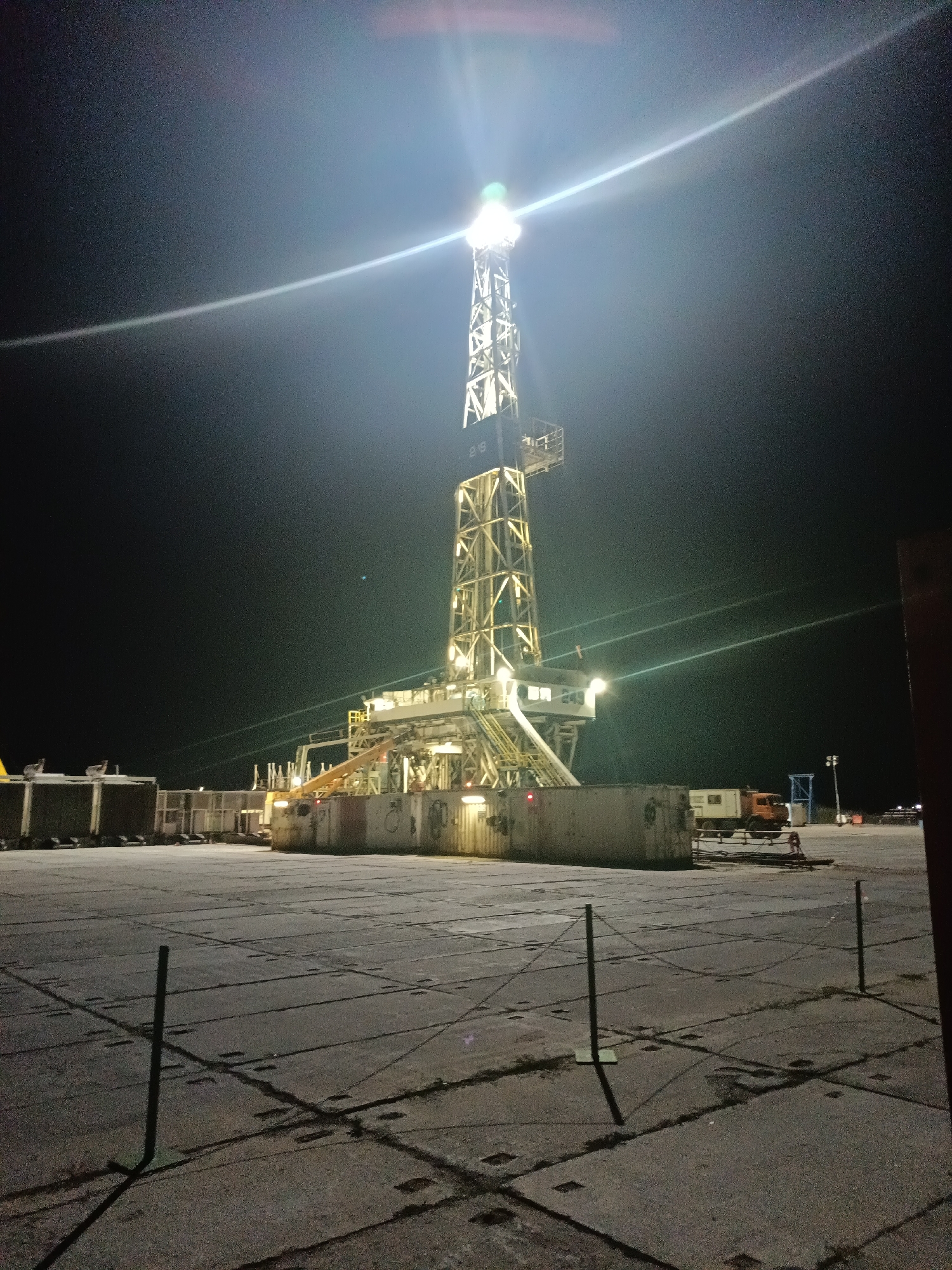 The image depicts a nighttime scene at an industrial site, likely an oil drilling operation, located in Пугачёв ауылдық округі, Kazakhstan. A towering drilling rig, illuminated by bright lights, dominates the center of the frame. The rig is a complex metal structure with multiple platforms and equipment. Surrounding the rig are several industrial containers, some appearing to be shipping containers. A truck is visible in the background, and a smaller portable building is parked near some of the containers. The ground is paved with concrete slabs, marked with lines indicating a grid pattern. The overall environment is dark, with the lighting from the rig and other sources creating strong contrasts and light flares. There are no people clearly visible in the image. The scene suggests ongoing industrial activity at night.