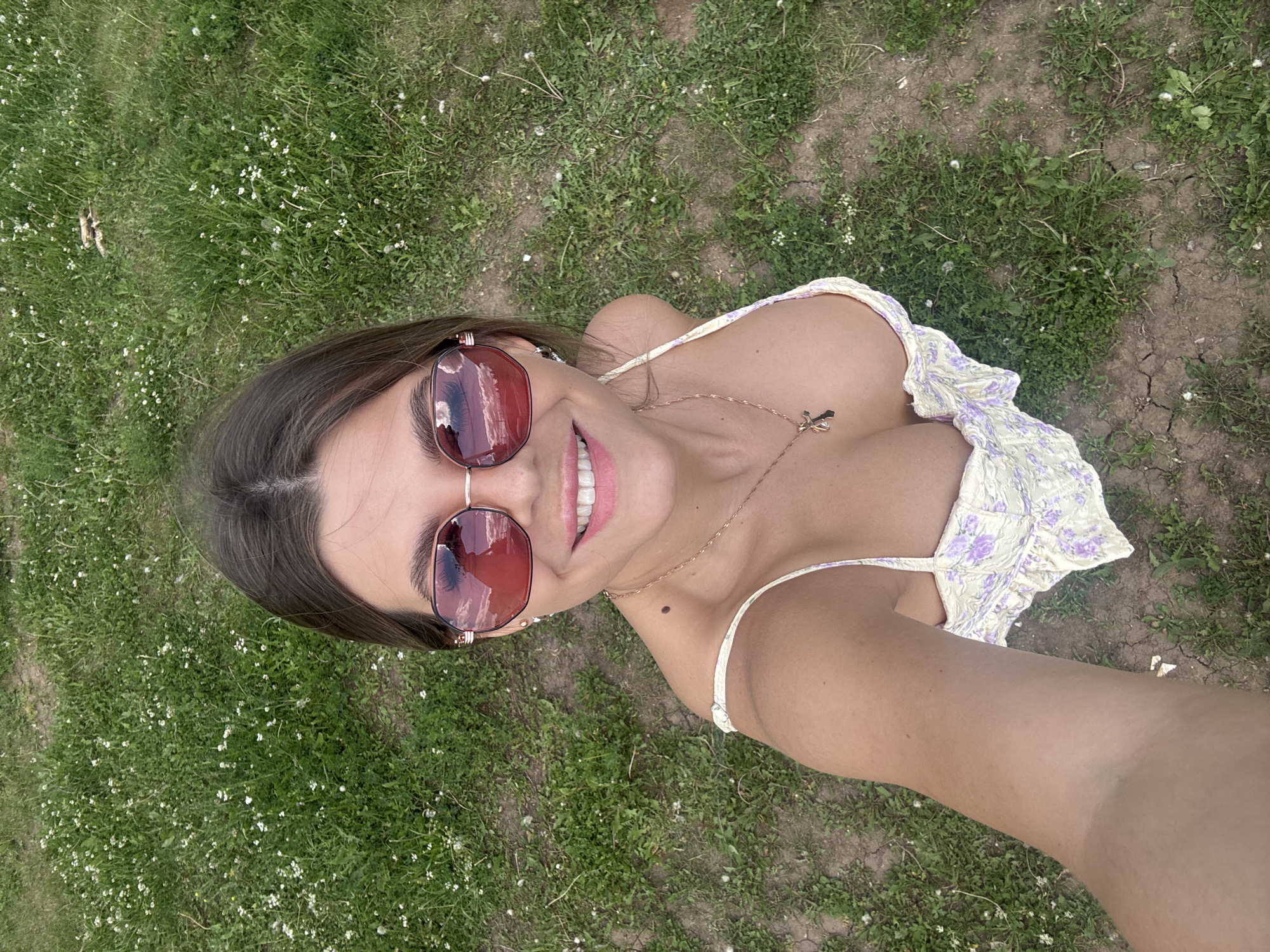 A young woman with dark hair, wearing rose-tinted sunglasses and a yellow and purple floral top, smiles at the camera while lying on her back in a grassy area. Her head is tilted back, and her hair is spread out around her. A delicate gold chain with a cross pendant rests on her chest. The background consists of green grass with scattered white wildflowers and patches of dry, cracked earth, suggesting an outdoor setting. The lighting indicates it is daytime, likely with a bright sun, given the reflections in her sunglasses. The overall mood appears relaxed and cheerful. The location is identified as Pastukhovo, Russia.