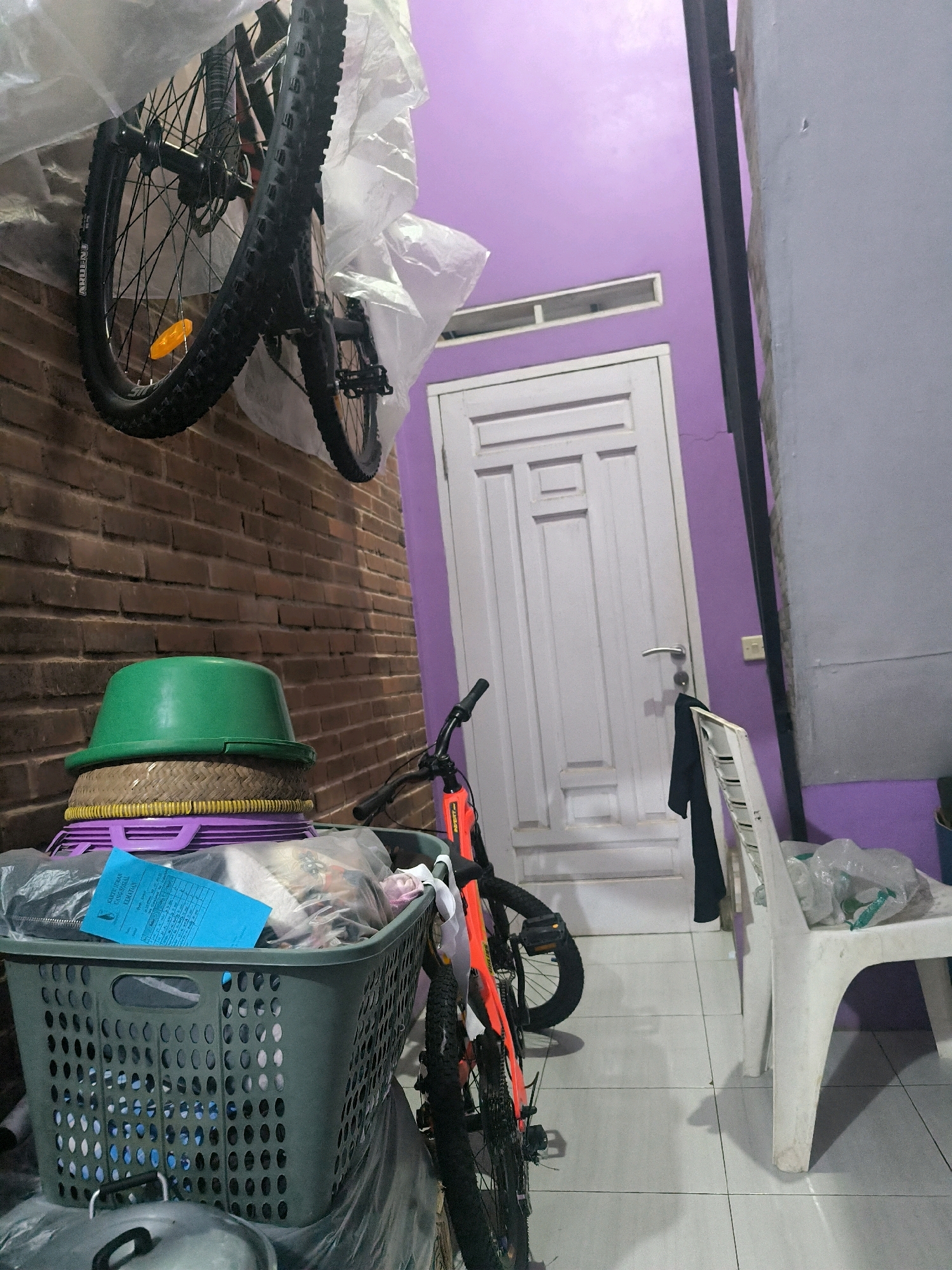 The image depicts a storage area, likely a porch or garage, with a variety of items. In the foreground, a green plastic laundry basket filled with various objects sits on top of other stacked items, including what appears to be a woven mat and a large green bowl. A blue document is placed on top of the contents of the basket. To the right of the basket, a bicycle with an orange frame is leaning against the wall. Another bicycle is suspended from the ceiling, partially wrapped in plastic sheeting, with its wheels visible against a brick wall. The background features a white door with paneling and a wall painted a light purple. A white plastic chair is positioned to the right of the door, with a dark garment draped over its back. The floor is tiled. The scene is indoors, with artificial lighting. The location is identified as Cilengkrang, Indonesia.