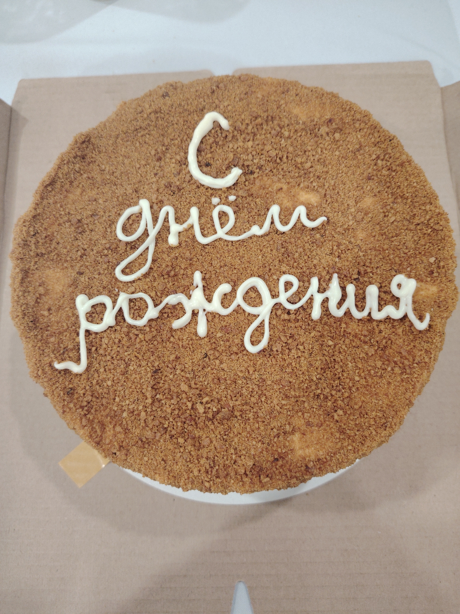 A close-up, overhead shot displays a round cake resting on a white plate within a brown cardboard box. The cake has a textured, light brown surface resembling crumb toppings. White frosting decorates the top, forming Cyrillic script, which translates to "Happy Birthday." The box, likely for transport or storage, is typical of pastry packaging. A beige plastic utensil is on the left side of the cake, which is presumably for serving. The image suggests a celebration event, possibly taking place in Sarapul, Russia.