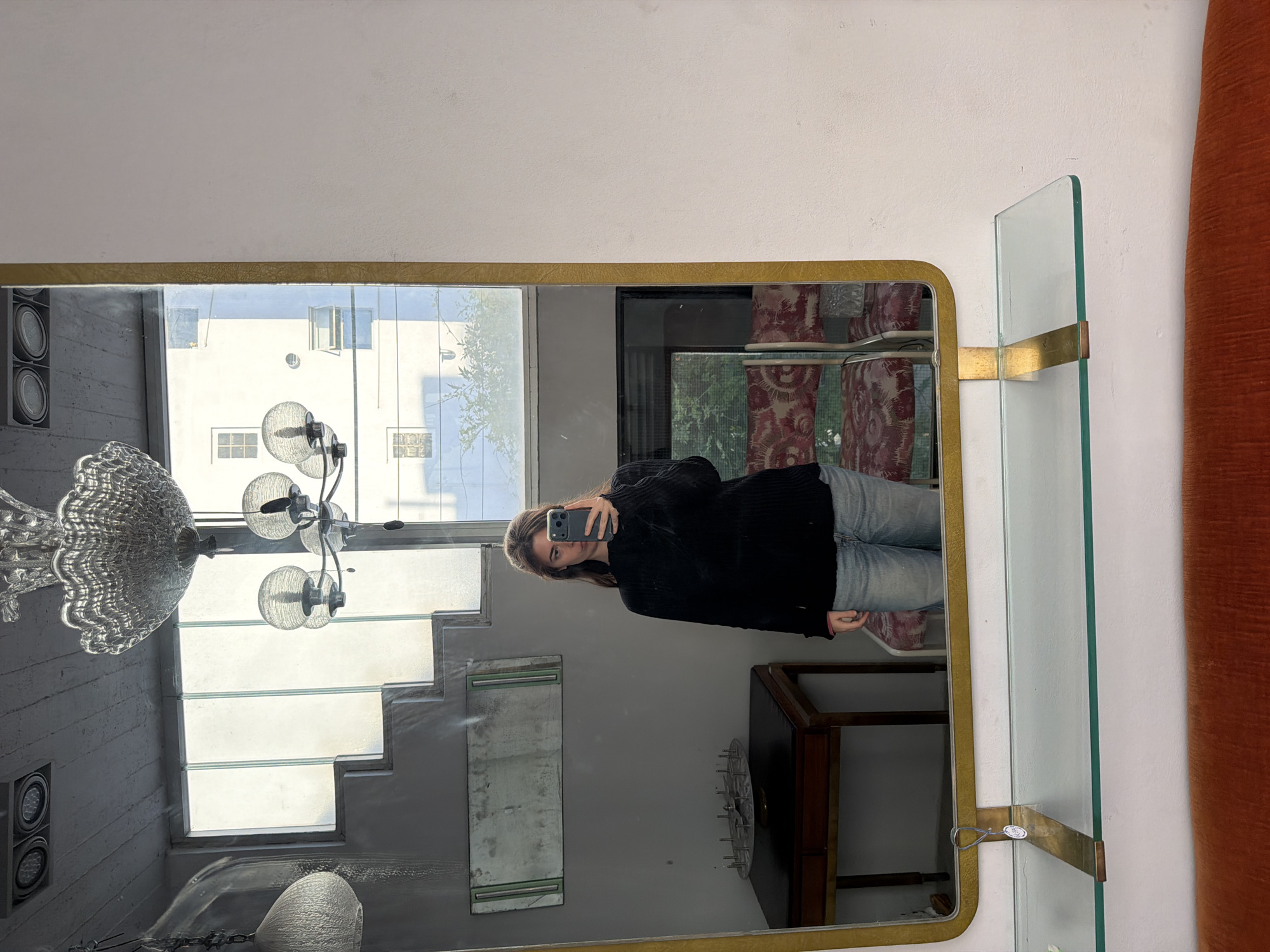 An indoor selfie taken in a large mirror with a gold-colored frame, showing a woman holding a smartphone. The woman, wearing a dark top and light-wash jeans, is reflected standing centrally in the mirror. To the right of the mirror, a glass shelf is attached with brass-colored fittings. The reflection reveals a diverse setting: to the left, a large, ornate clear glass chandelier hangs, and a set of stairs with a grey wall is visible behind it. Outside the windows, reflected in the mirror's upper left, is a white building with multiple windows, some open, and hints of trees. Another light fixture with spherical glass shades is also reflected in the upper left. To the right of the woman, reflected patterns of red and white fabric (possibly a curtain or upholstery) are visible. A dark wooden table or cabinet with a decorative metal plate is reflected below the woman. The overall scene suggests a space with a mix of vintage and classic elements, possibly a home or a shop. The setting is Buenos Aires, Argentina. A small, white tag with faint, unreadable text (possibly a price or product tag) hangs from the brass support of the glass shelf. The time of day appears to be daytime due to the natural light.