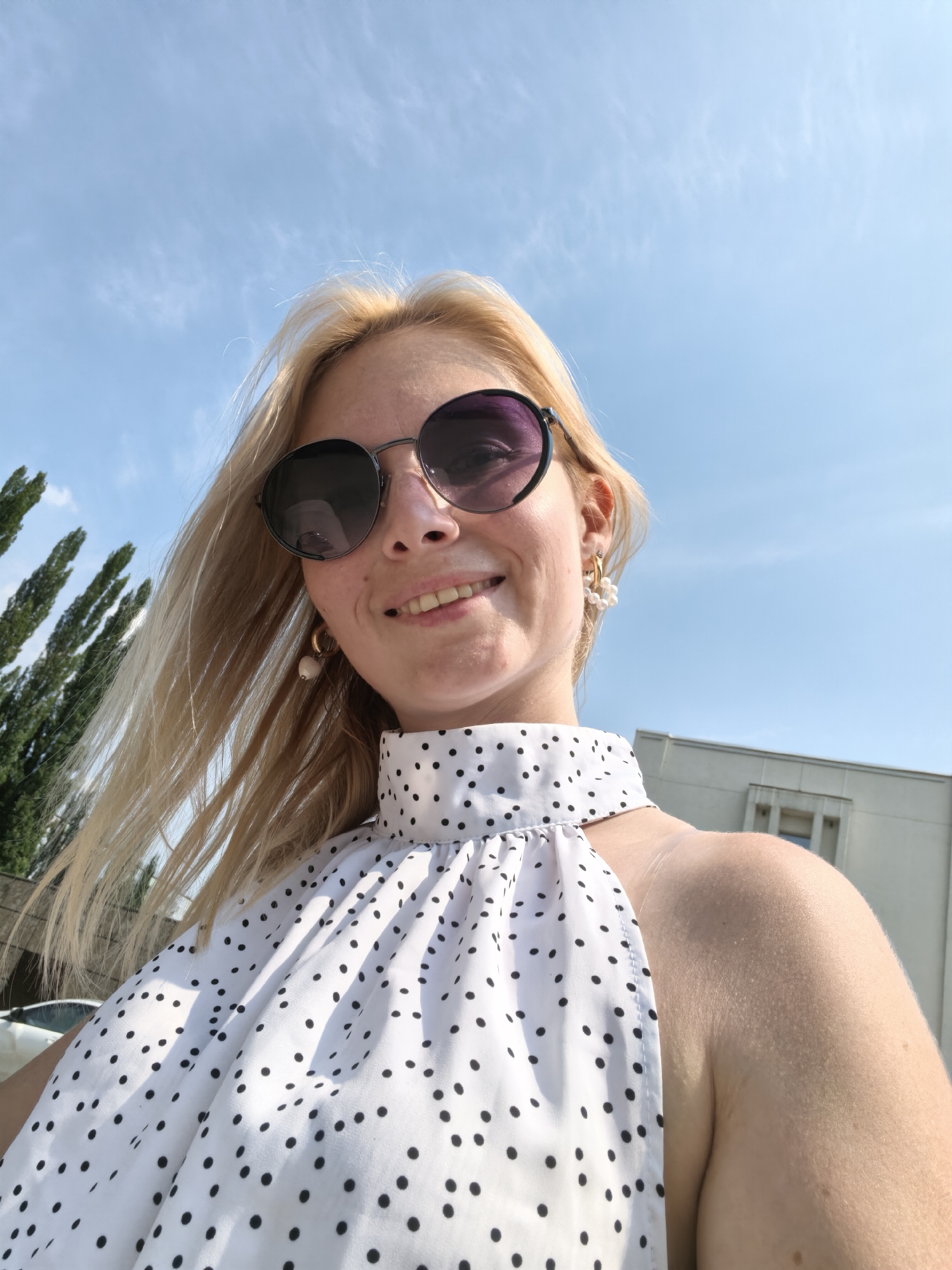 A blonde woman wearing sunglasses and a white polka-dot dress is taking a selfie outdoors. She is smiling, and her hair is blowing in the wind. The background shows a clear blue sky with some wispy clouds, trees, and a building. The image appears to be taken on a sunny day. The specific location within the city of Sterlitamak, Russia, cannot be determined from the visual information provided.