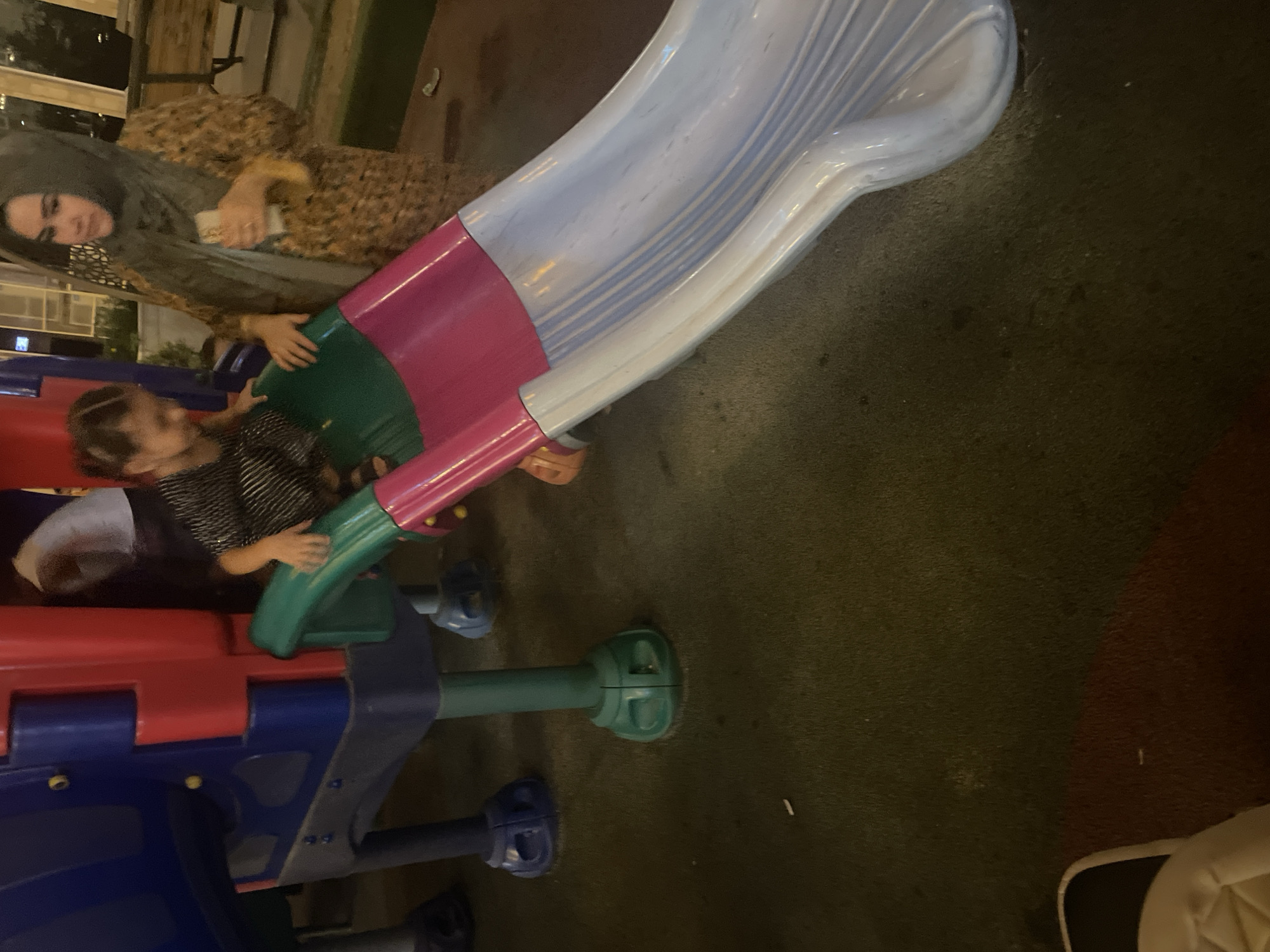 The image depicts a playground scene, likely in Dushanbe, Tajikistan, featuring a colorful slide and two individuals. A young girl, dressed in a black and white striped shirt, is positioned on the green and pink upper section of the slide, her hands gripping the sides as if about to descend. Beside her, on the right, a woman wearing a hijab and a patterned dress looks towards the girl, her right hand holding what appears to be a mobile phone. The playground equipment is made of brightly colored plastic, with blue and red components forming the structure. The slide itself is a curved surface, transitioning from pink at the top to a pale blue with white stripes as it descends. The ground beneath the slide is covered in a dark, textured material, possibly artificial turf or rubberized playground surfacing, with some scattered debris. In the background, other playground structures are partially visible, along with hints of urban surroundings, including lighting fixtures and possibly buildings, suggesting it is an outdoor public space. The overall lighting suggests it may be evening or a dimly lit indoor space.