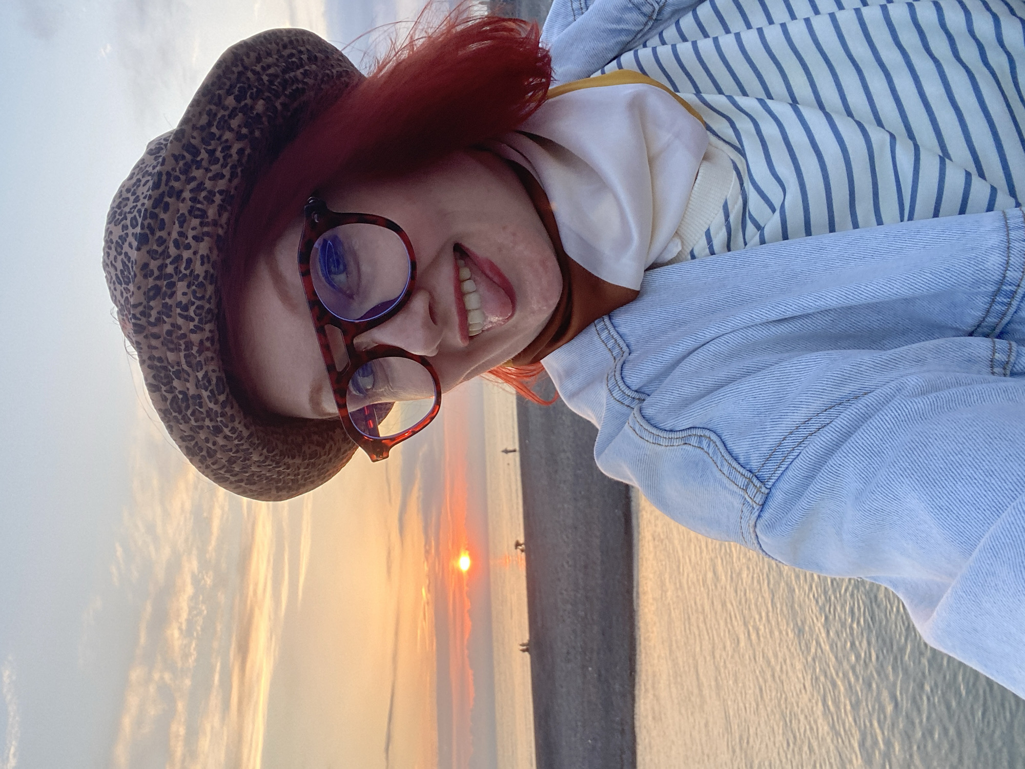 A woman with red hair and glasses is posing for a selfie in Sochi, Russia. She wears a leopard-print hat, a blue striped shirt, and a denim jacket. Her tongue is sticking out. The sunset is visible over the Black Sea, and some people are walking along the beach in the background. The scene suggests a casual, touristy moment.