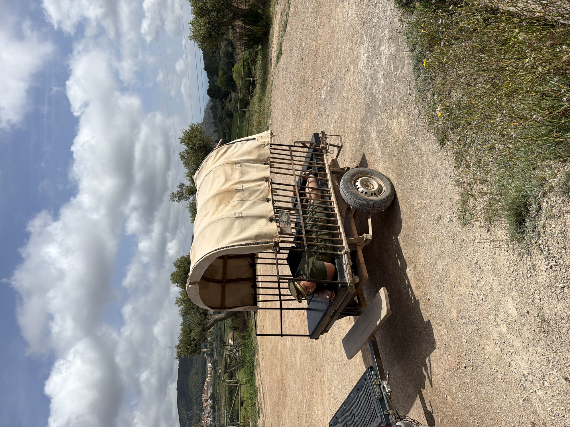 A man wearing a khaki uniform lies inside of a wagon-like trailer with a cream-colored canvas top and a steel cage frame, which is hitched to the back of a dark-colored pickup truck. The trailer has one tire on the visible side. The scene is outdoors, on a dirt road with vegetation along the edges. In the background, there is a view of rolling hills and a distant village, consistent with the landscape around Bonastre, Spain. The sky is partly cloudy.