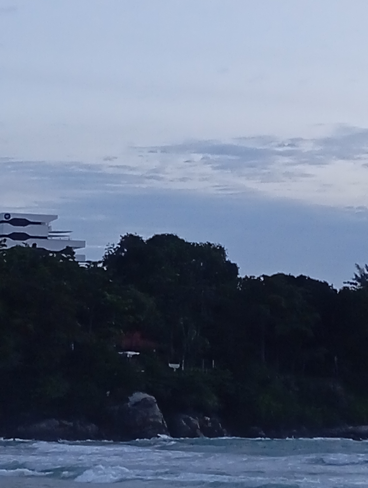 The image depicts a coastal scene in Karon, Thailand, likely taken from the water. In the upper portion of the frame, the sky is overcast with muted shades of blue and gray, with faint cloud formations visible. Below the sky, a dark green, densely forested hillside dominates the mid-ground. Perched atop the hillside, a modern white building with distinctive horizontal bands and curved sections is partially visible. The building appears to be a resort or hotel, given its scale and design. The foreground is occupied by the sea, with rough, choppy water in shades of blue and white. Small waves are breaking, indicating a somewhat turbulent sea. The overall lighting suggests it is either early morning or late evening, with soft, diffused light. No people are visible in the frame, and no specific actions or interactions are captured. The scene is devoid of any identifiable landmarks that would pinpoint the exact location within Karon, other than the general coastal environment.