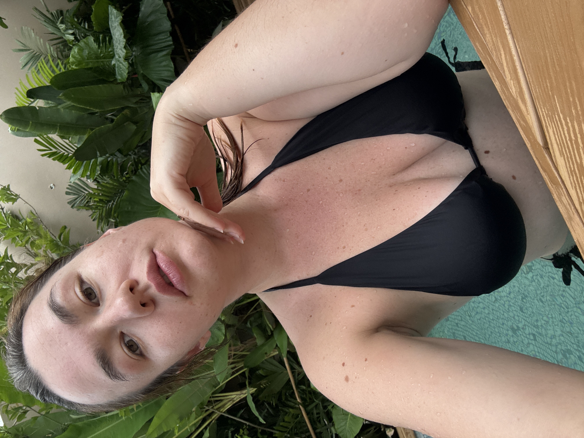 A woman in a black bikini poses for a selfie in a swimming pool, with lush green foliage in the background. She has her arm raised, with her hand resting on her chin. The image appears to have been taken outdoors, possibly in a tropical setting. The water in the pool is a bright turquoise color, and there are wooden planks visible at the edge of the pool. The woman has her hair wet, suggesting she has recently been in the water. Her skin is speckled with water droplets. The overall impression is one of a relaxed, vacation-like scene.
