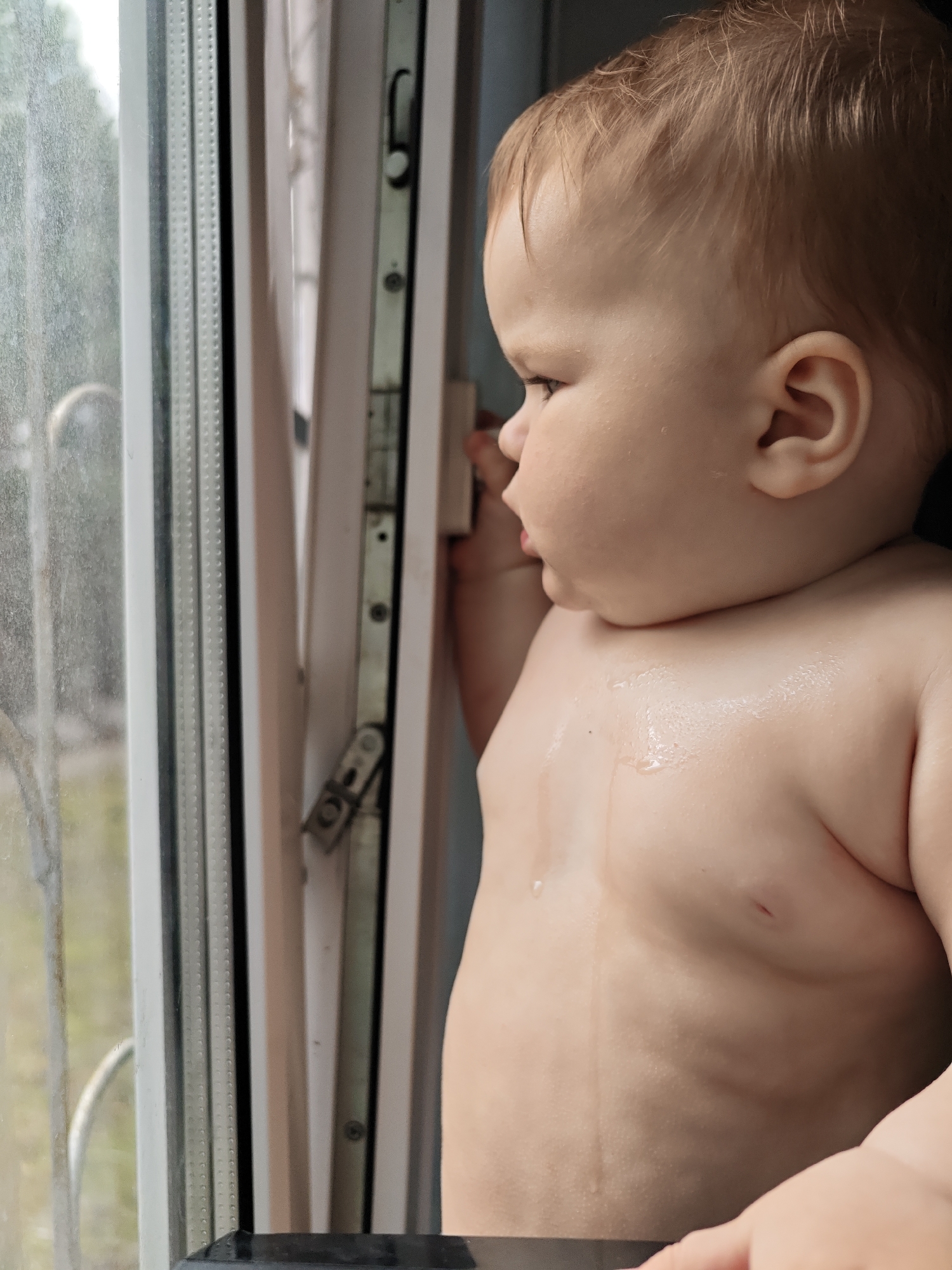 A baby with light red hair stands topless facing a window, looking outside in Chaikovsky, Russia. The window is open and has a metal mechanism. The baby is holding onto the window frame with one hand and is mostly facing left. Water droplets are visible on the baby's chest. The scene is illuminated by natural light.