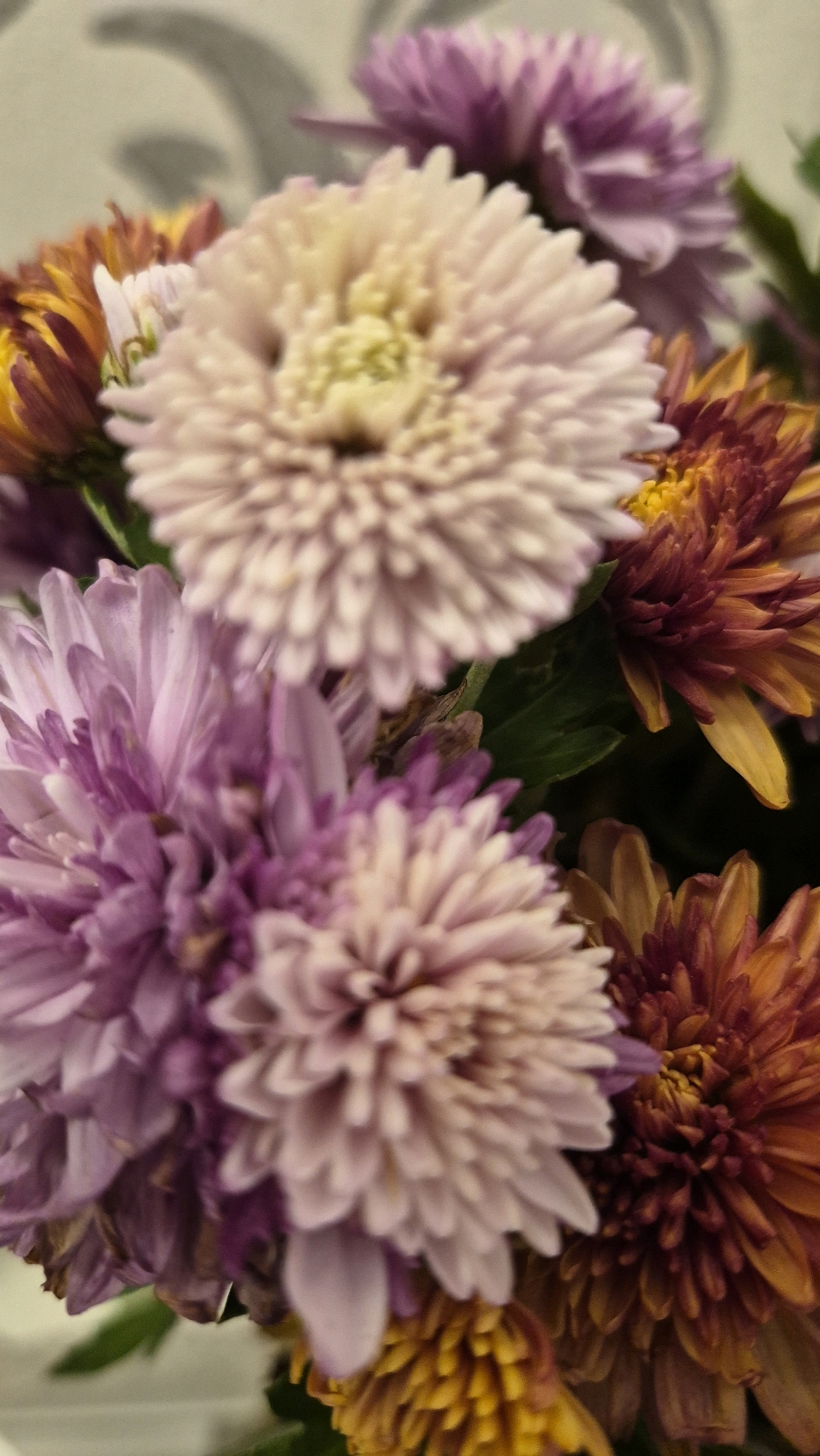 A close-up, vertical shot shows a bouquet of chrysanthemums in various shades of pink, purple, and orange. The flowers are tightly packed, with their layered petals creating a textured appearance. The background is blurred and appears to be a light-colored wall with an abstract, grey pattern. The lighting is soft, highlighting the intricate details of the flower petals. The scene appears to be indoors, possibly in a home or flower shop, and there are no people or animals present. The image is entirely focused on the floral arrangement.
