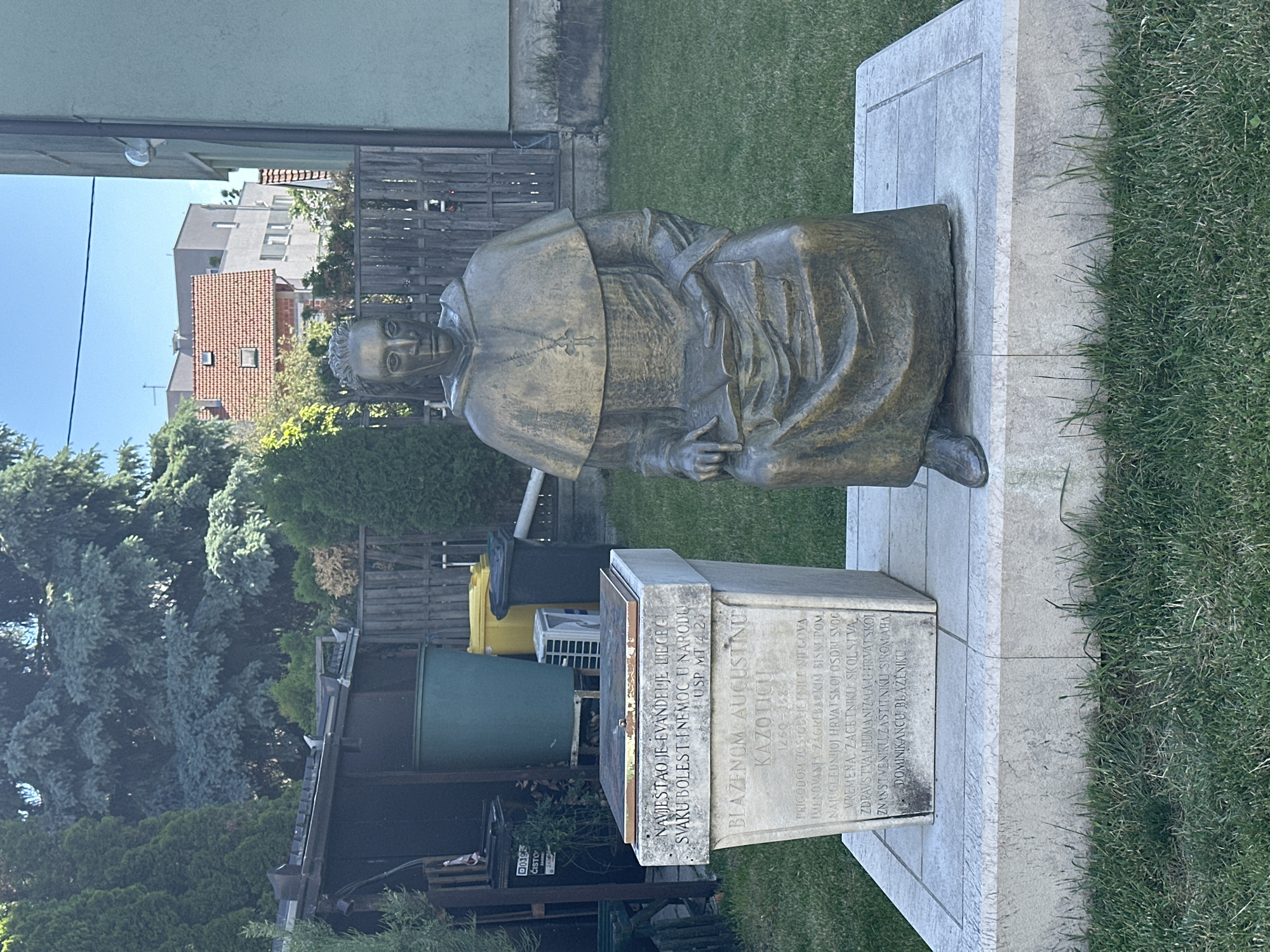 Here's a detailed description of the image:
The image features a bronze statue of Blessed Augustin Kazotic, a Croatian Dominican friar who was the Bishop of Zagreb in the 13th and 14th centuries. The statue depicts him in a seated position, wearing a bishop's robes and head covering. The statue is mounted on a stone base with inscriptions. The inscription on the monument states that he was "the most distinguished of Croatian people."
The statue is located outdoors, with a building and trees in the background. The statue is standing on a stone base sitting on the ground in a grassy yard. A large olive-green rain barrel and a pair of refuse containers are in the background. The sky is clear and blue. The location of the statue is likely in the city of Zagreb, Croatia, though there are no visual elements in the image that confirm the exact location within Zagreb.