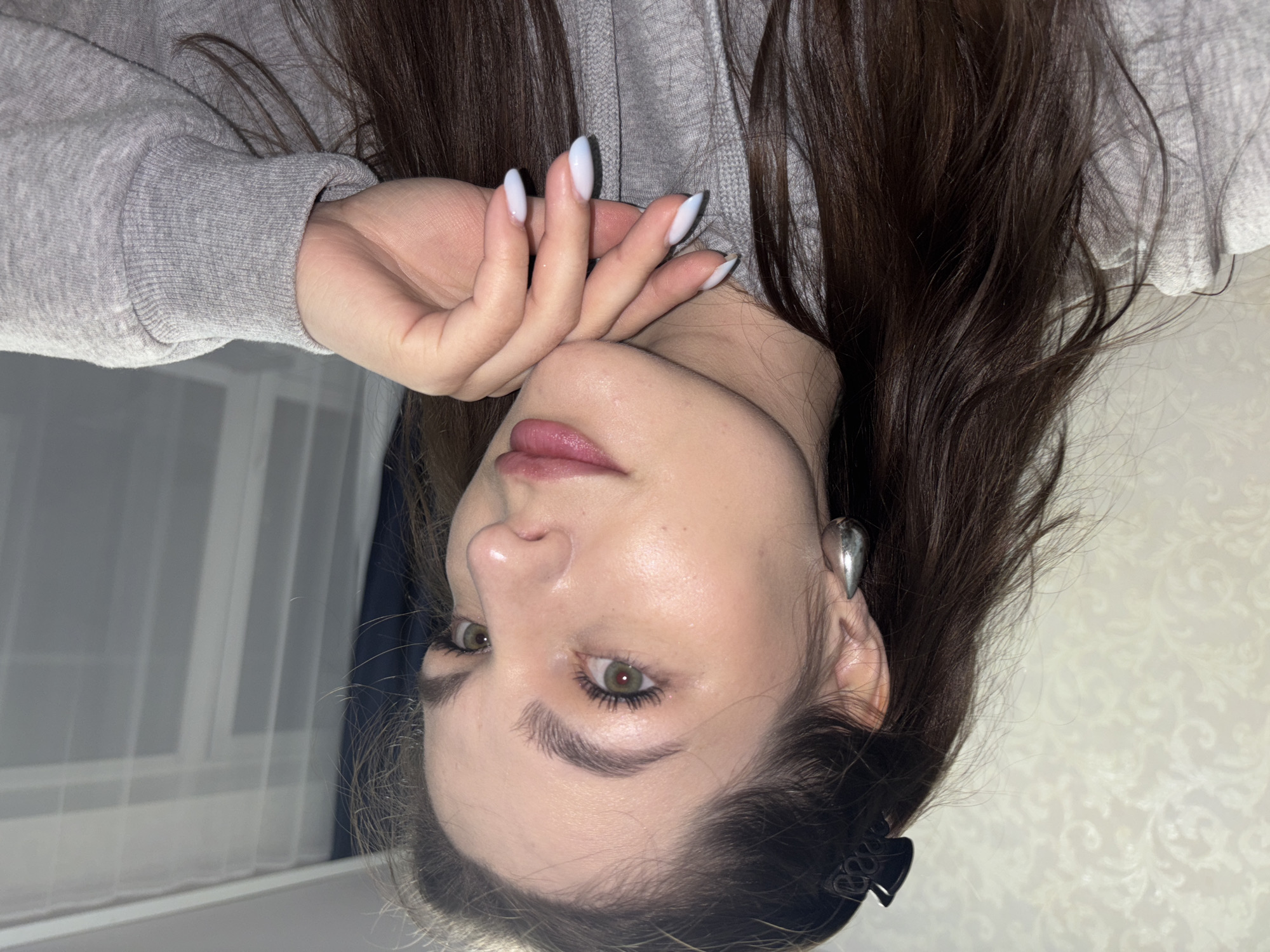 A young woman is depicted indoors, likely in a residential setting. She has light skin, brown hair, and is wearing a light gray hoodie. Her nails are painted a light color. A silver, teardrop-shaped earring is visible on her left ear. Her hair is styled and dark brown. A black hair clip is visible on the lower right part of the photo. The background includes a curtain and patterned wallpaper. There is insufficient information to determine if the photo was taken in Chaikovsky, Russia, or to ascertain any specific actions or interactions.