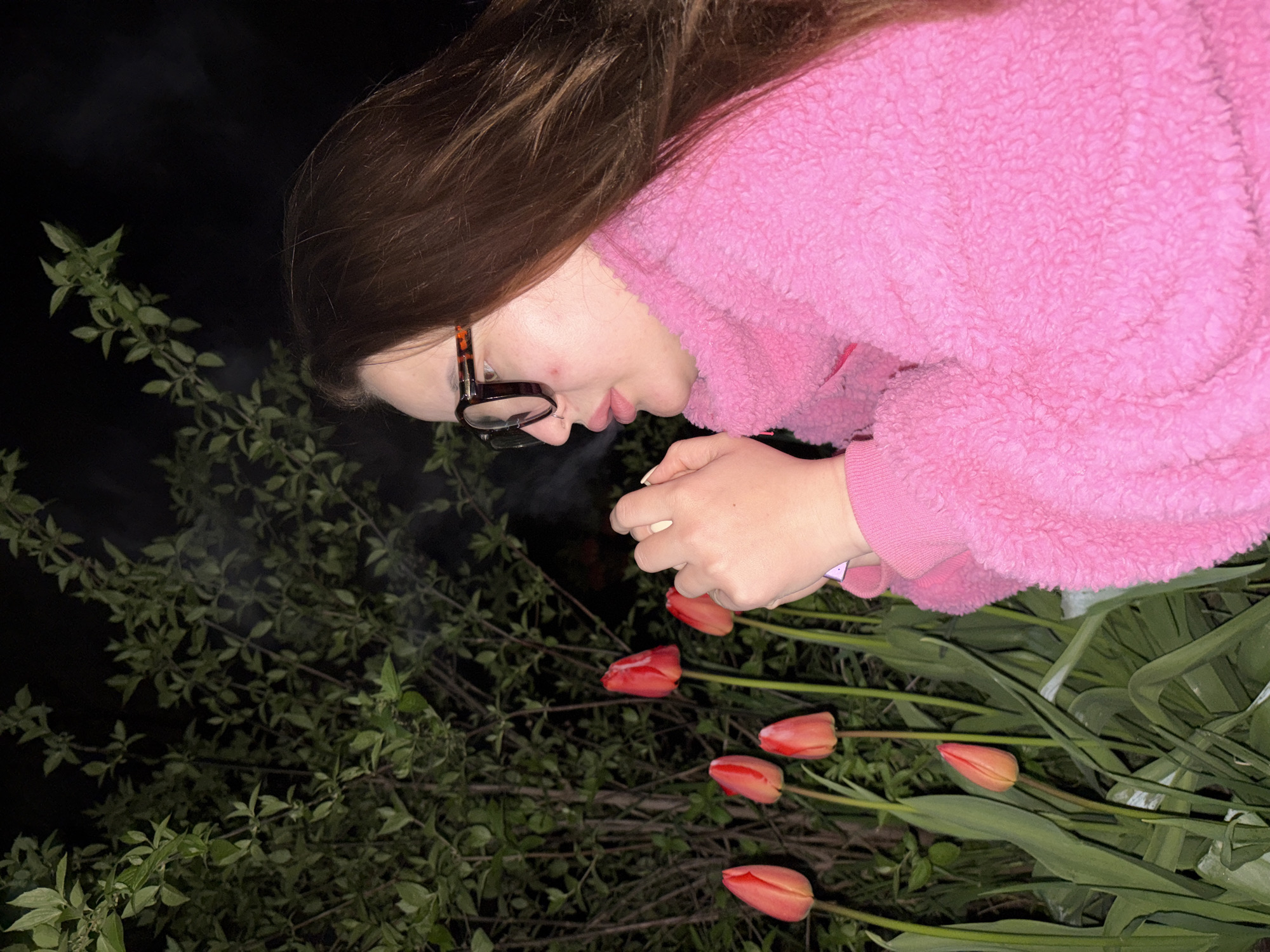 A woman wearing glasses and a pink fuzzy jacket is holding a small, white object over a patch of green plants and a row of red tulips. The setting appears to be outdoors at night, given the dark background and the presence of the plants. The specific location is not discernible from the image.