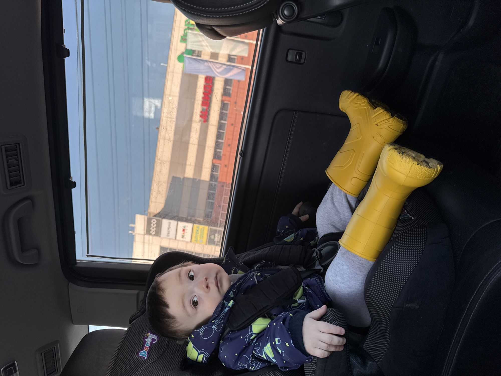 A child in a car seat wearing a patterned jacket and yellow rubber boots is visible in the image. Through the car window, the "J.BMAE" building and "ECCO" store can be seen, indicating the location is likely Izhevsk, Russia.