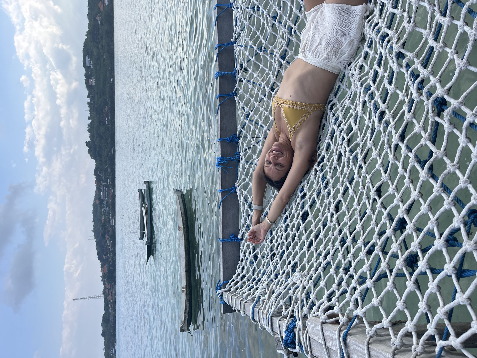 A woman with dark hair is lying on a white net hammock suspended over water. She is wearing a yellow crocheted bikini top and white linen shorts. Her arms are stretched above her head, and she is smiling at the camera. The hammock is attached to wooden beams. In the background, there are several small boats on the water. Beyond the water, there is a coastline with trees and buildings, including a prominent telecommunications tower. The sky is partly cloudy. This scene is likely located in Nusa Penida, Indonesia, an island known for its scenic coastlines and ocean activities.