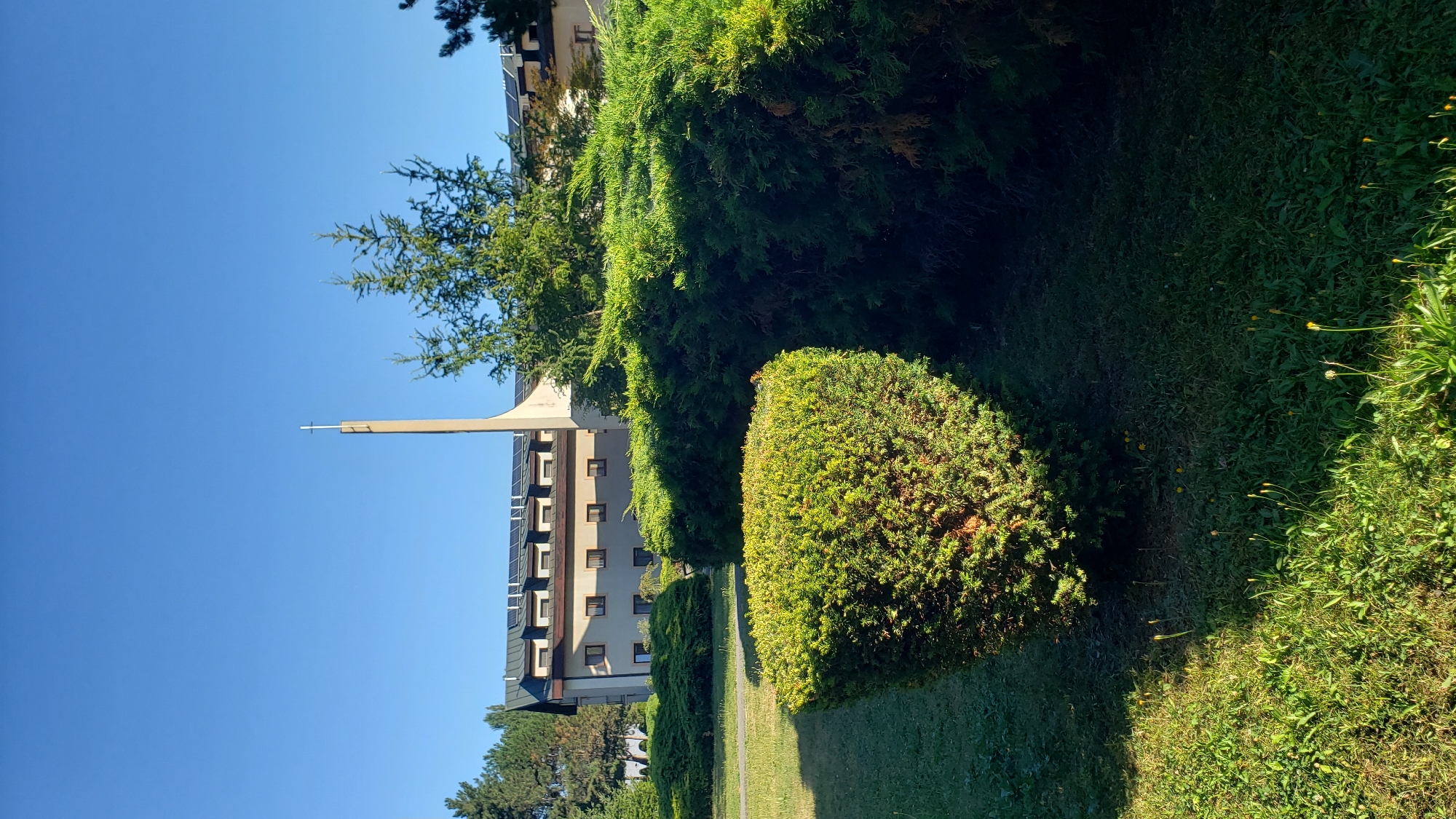 The image depicts the exterior of a church or religious building in Prešov, Slovakia, under a clear blue sky. In the foreground, there is a well-manicured lawn with a prominent, rounded evergreen bush. Behind this, a dense hedge forms a natural barrier. The main structure is a multi-story building with a dark roof featuring multiple dormer windows, some of which appear to have solar panels installed. A tall, slender tower, topped with a cross, rises from the side of the building. The architecture suggests a modern design with a religious function. The overall scene is tranquil and bathed in sunlight, highlighting the green foliage and the building's structure.