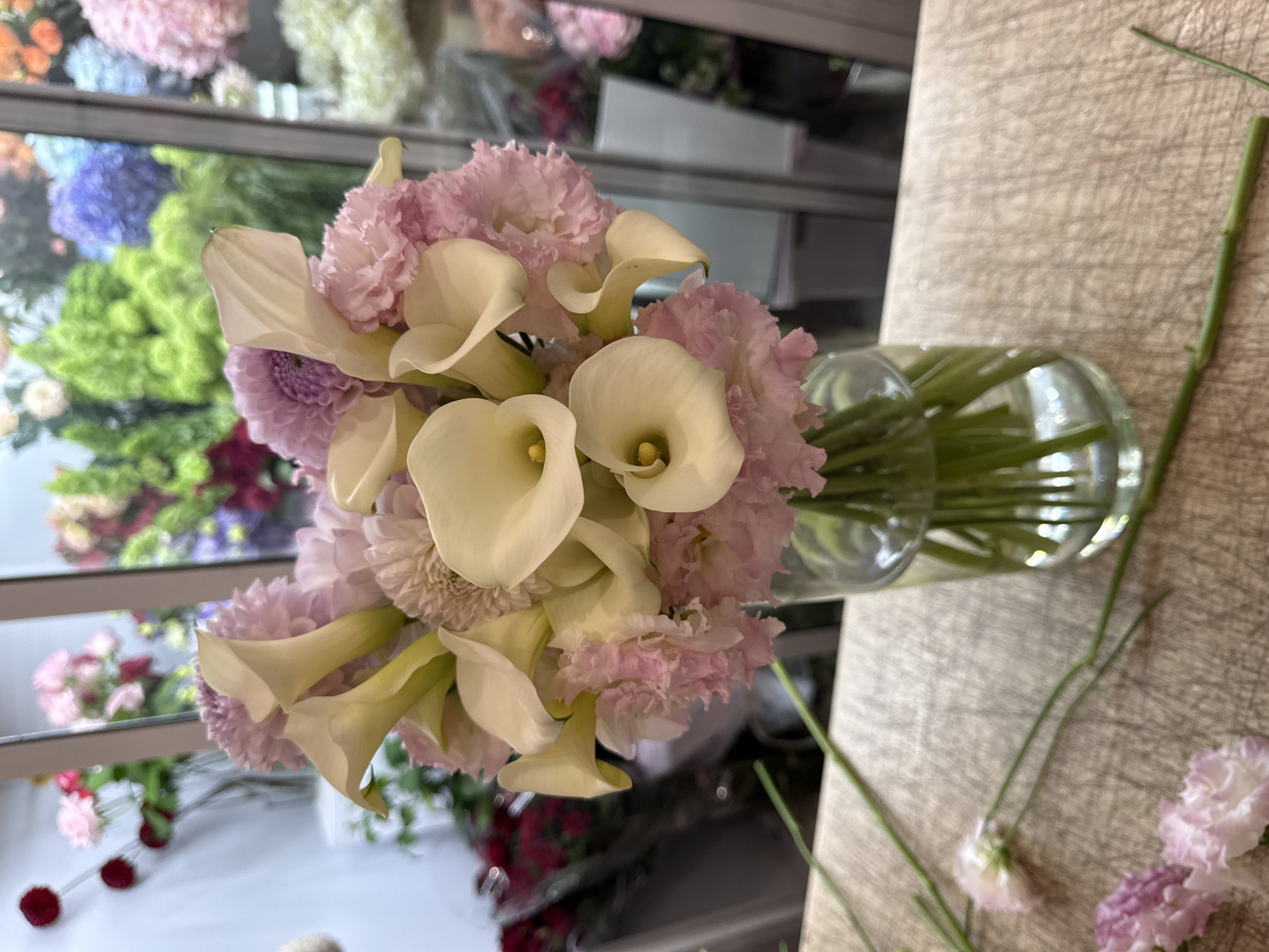 A close-up, eye-level shot shows a bouquet of flowers in a clear glass vase. The bouquet is composed of white calla lilies with yellow centers, pale pink carnations, and light purple dahlias. The flowers are arranged tightly together, with their stems submerged in water within the vase. The vase is positioned on a light brown, textured surface that appears to be a wooden table with visible grain and scratches. In the background, a blurred display of various colorful flowers in vases and buckets is visible through a window or glass partition. The lighting is bright, highlighting the details of the flowers and the vase. The image does not contain any people or show any actions or interactions beyond the arrangement of the flowers. The location is not specified in the image or the provided information.