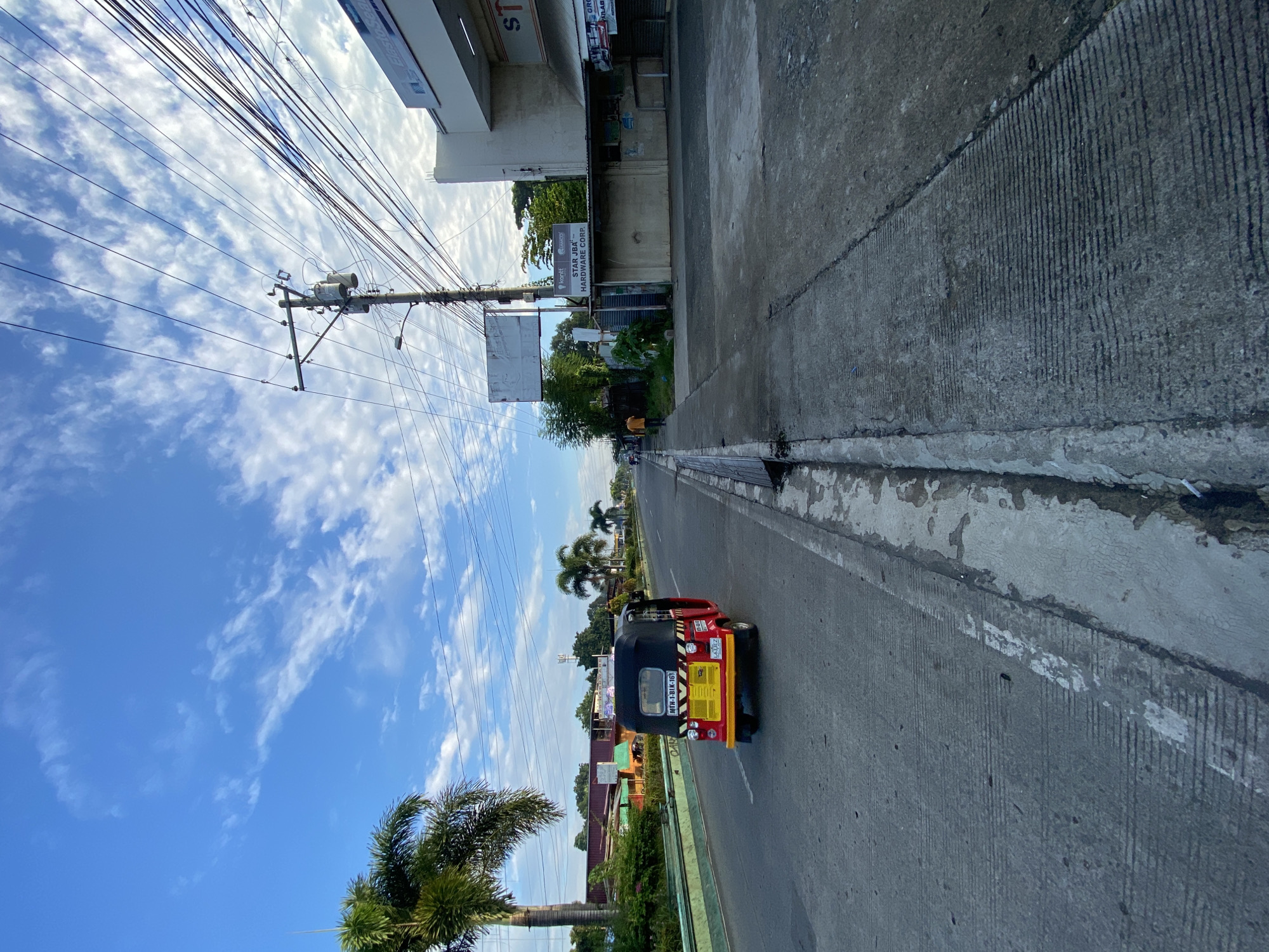 The image depicts a street scene in Davao City, Philippines, captured under a bright, partly cloudy blue sky. A red and yellow motorized tricycle, identified by the license plate "MTH-1-BIK-161", is driving away from the viewer on the right lane of a paved road. The road is lined with a concrete sidewalk on the right and a grassy median with a curb on the left.
To the right of the road, several buildings are visible, including a beige structure with a sign that reads "STAR JBA HARDWARE CORP." A utility pole with a transformer and numerous overhead wires stands prominently in front of the buildings. Further down the road, more structures and greenery are present. On the left side of the road, a palm tree and a green barrier are visible, suggesting a tropical environment. Another person, wearing an orange shirt, is walking on the sidewalk in the distance. The overall impression is a typical urban or suburban street in the Philippines during daylight hours.