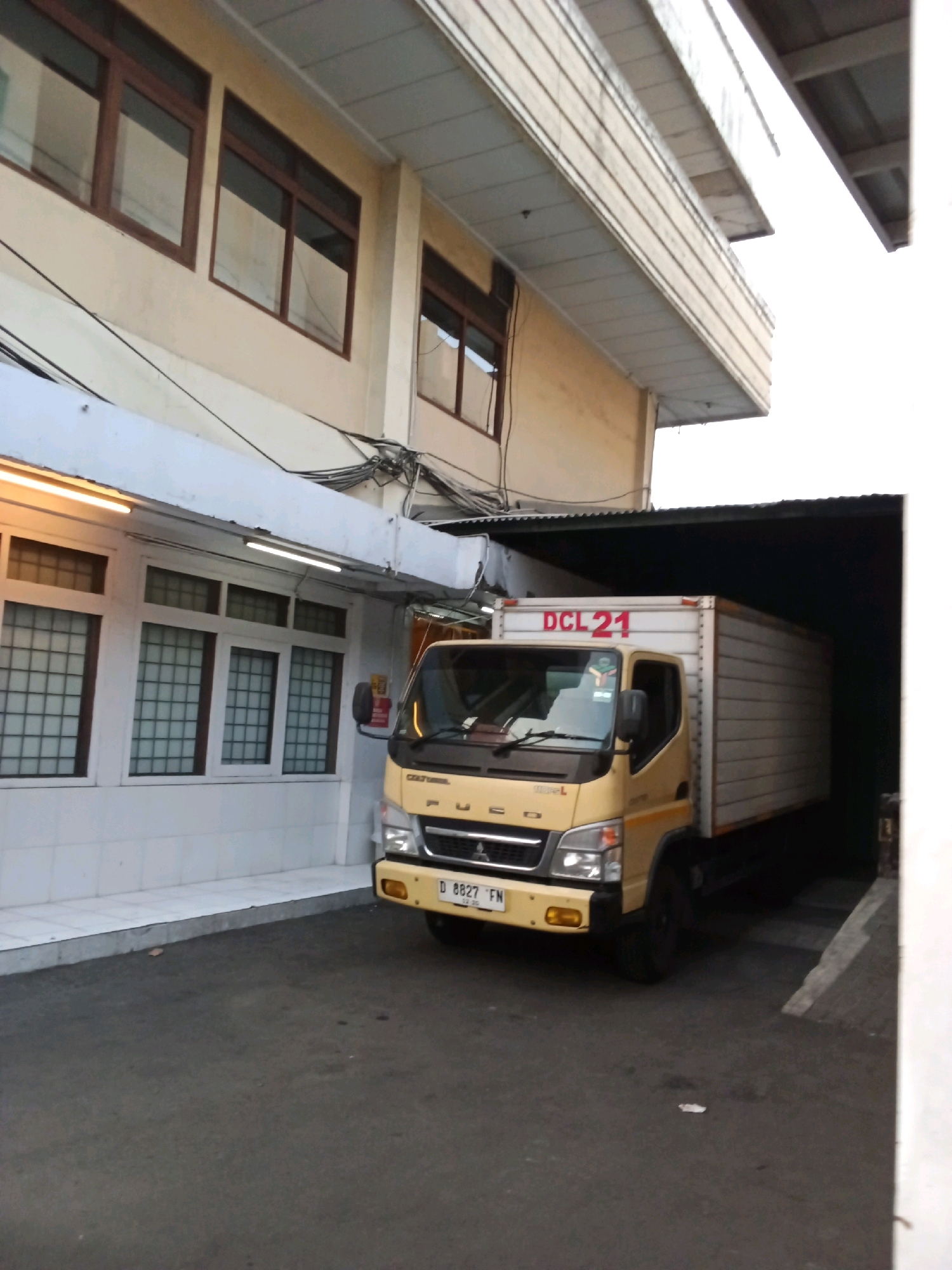 A yellow truck with "DCL 21" written on its side is parked under a covered walkway of a building. The truck has a license plate that reads "D 8827 FN". The building appears to be an office or industrial building, with several windows visible on the upper floors. The ground is paved with asphalt. The scene suggests a loading or unloading operation at a facility in Bandung City, Indonesia.