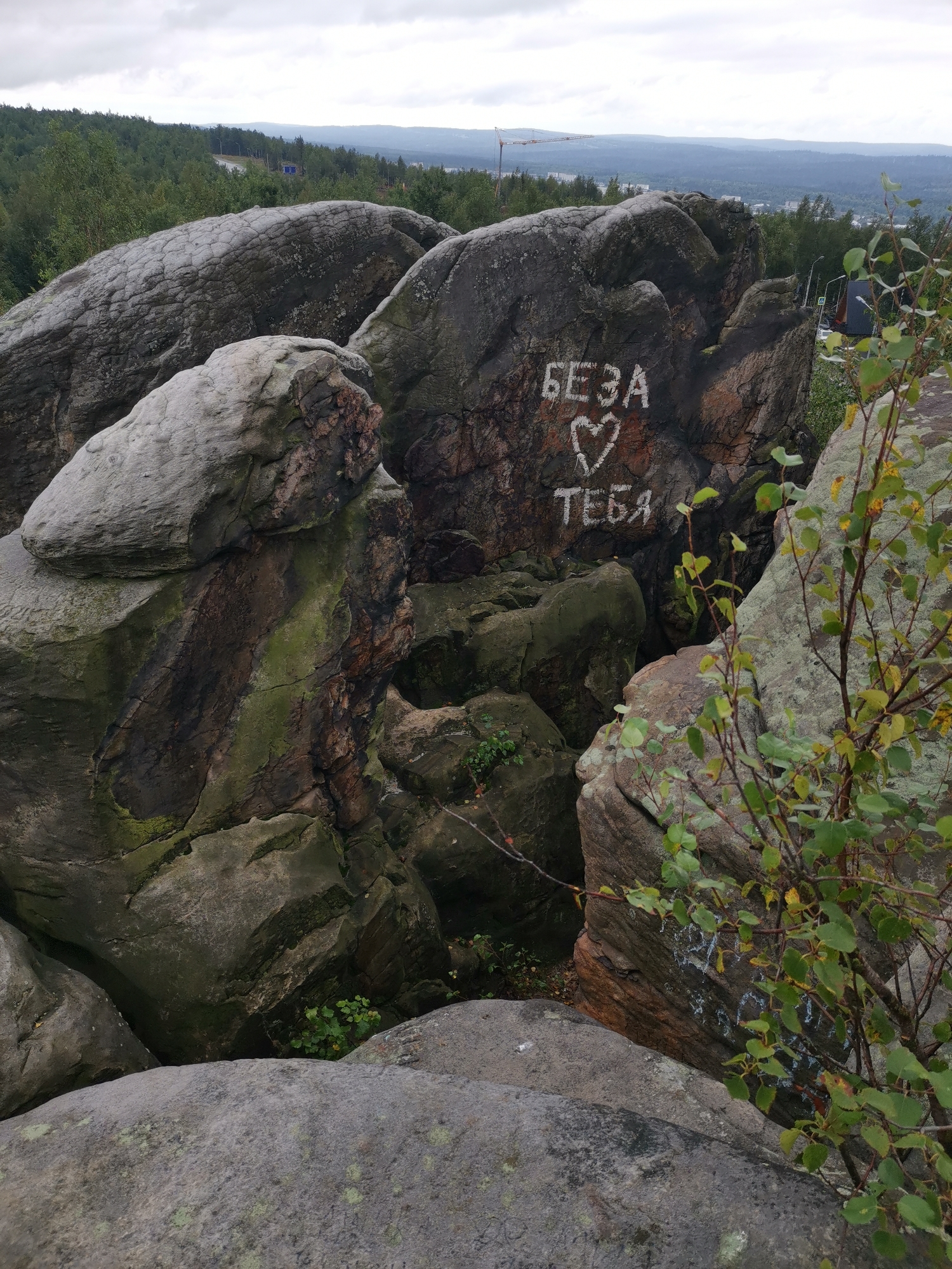 The image shows a rocky outcrop with graffiti on one of the rocks. The graffiti, written in white paint, reads "БЕЗА ♡ ТЕБЯ" which translates to "Beza, I love you." The rocks are large and irregularly shaped, with some showing signs of moss and lichen growth. In the background, a dense forest stretches towards a hazy horizon, with distant hills and a faint urban landscape visible. The sky is overcast, suggesting a cloudy day. The scene appears to be an outdoor natural setting, possibly a viewpoint or a recreational area. This location is identified as part of the local natural park in Gubakha, Russia, a region known for its karst formations and scenic beauty.