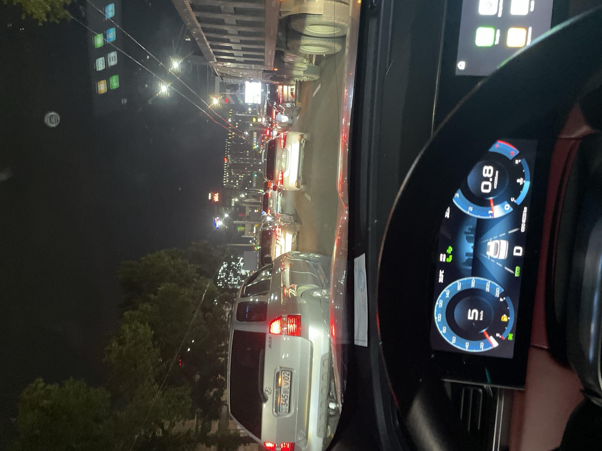 The image depicts a nighttime view from the driver's perspective of a car stopped in heavy traffic on a city street. The road is lined with trees and streetlights, and illuminated buildings are visible in the background. Several cars, including SUVs and sedans, are in front of the vehicle, some with their brake lights illuminated. A large dump truck is visible to the right of the frame. The car's digital dashboard is visible, showing a speed of 5 km/h and other vehicle information. The license plate of the silver SUV in front reads "345BLV02". The scene is consistent with typical evening traffic congestion in a large city.