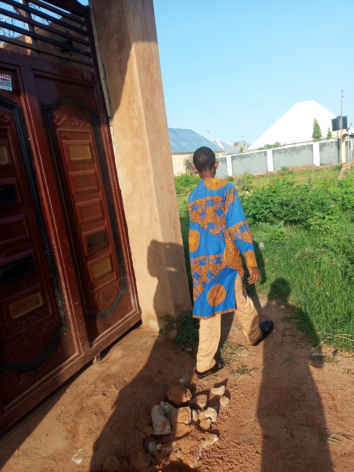 A young boy wearing a blue and orange patterned tunic and beige pants walks away from a large, ornate brown metal gate. The gate has decorative panels and is partially open, revealing a sliver of a darker interior. To the right of the gate is a rough, beige-colored pillar, casting a long shadow across the sandy ground. Beyond the pillar, a patch of green grass and dense foliage stretches towards a low wall. In the background, several buildings with corrugated metal roofs and a large, white, pyramid-shaped structure are visible under a clear blue sky. The scene appears to be an outdoor residential area in Tempure, Nigeria.