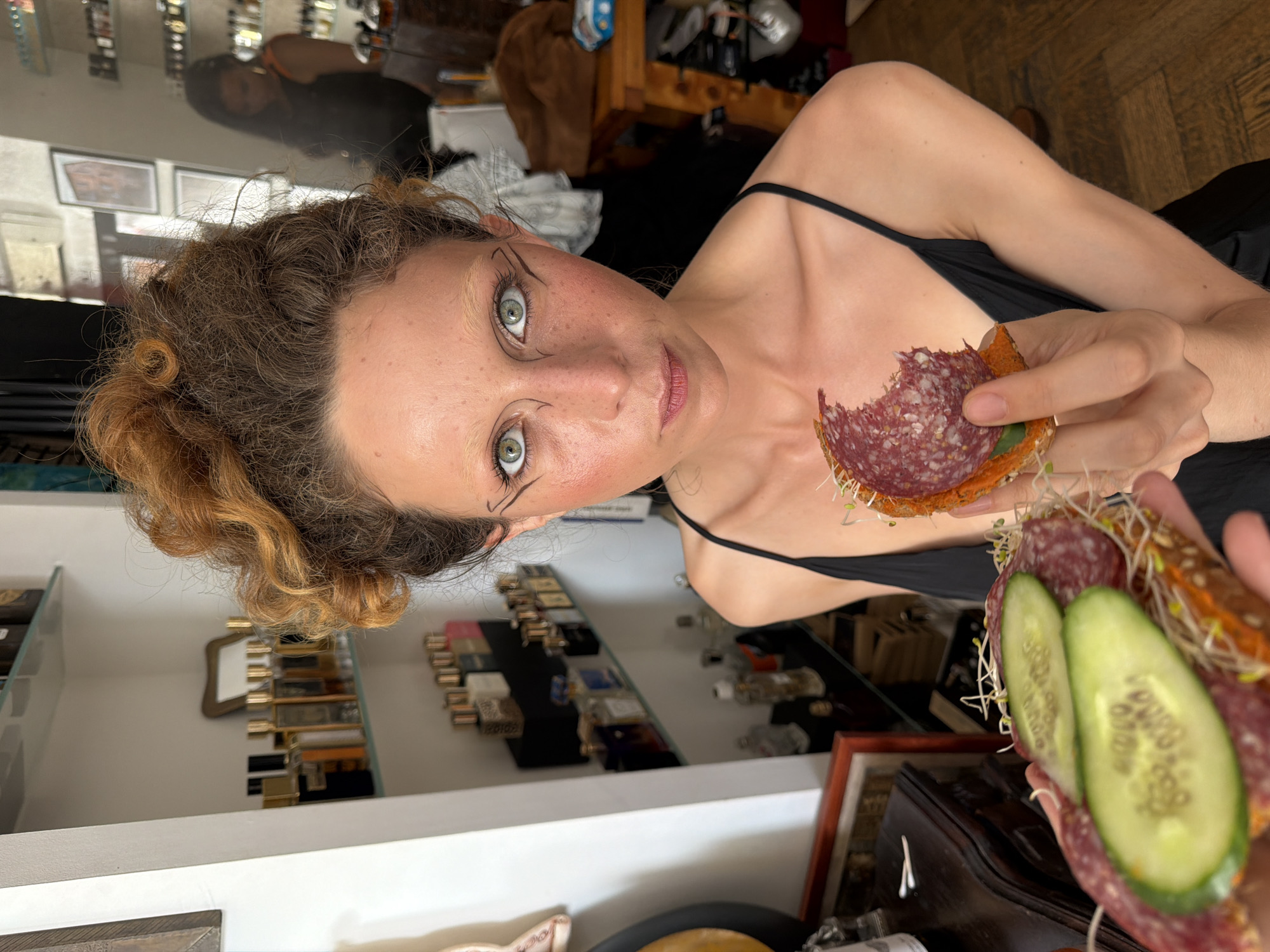 A woman with curly, auburn hair is holding two open-faced sandwiches. She is wearing a black camisole and has dramatic eye makeup. Her gaze is directed at the camera. The background suggests an indoor setting, possibly a shop or a home, with shelves stocked with bottles and framed artwork. One sandwich has slices of cucumber and salami with sprouts on what appears to be rye bread. The other sandwich, which she is biting into, also has salami and sprouts on bread. The scene is well-lit, and the focus is on the woman and the sandwiches. The floor is a wooden parquet. The woman's makeup and hairstyle suggest an artistic or theatrical presentation. The bottles on the shelves could be perfumes, oils, or other boutique products. The overall atmosphere is intimate and personal.