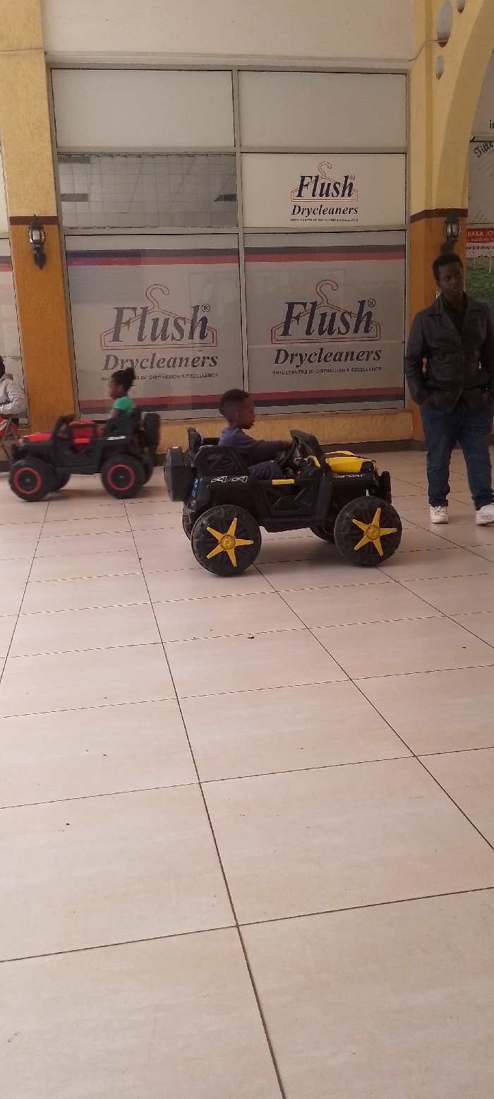 The image displays an indoor commercial area, likely a shopping mall corridor, located in Ngong ward, Kenya. The floor is composed of large, light-colored square tiles. The background features a storefront with multiple glass panels, prominently displaying "Flush Drycleaners" branding, including a hanger logo and the slogan "DRYCLEANERS OF DISTINCTION & EXCELLENCE." Structural columns with a textured yellowish-brown finish are visible; one on the left contains a dark sconce-style light fixture, and another on the right exhibits decorative arch cutouts.
Two young children, estimated to be between 3-5 years old, are operating electric ride-on toy vehicles. In the foreground, a child wearing dark clothing is seated in a black ride-on car with yellow accents and yellow star-spoked wheels, gripping the steering wheel and facing towards the right side of the frame. Further back on the left, another child, wearing a green top, is seated in a red and black ride-on car, positioned with their back to the camera. An adult male stands upright to the right, in the background, observing the scene. He is dressed in a dark jacket, blue jeans, and white sneakers, with his hands placed in his pockets.