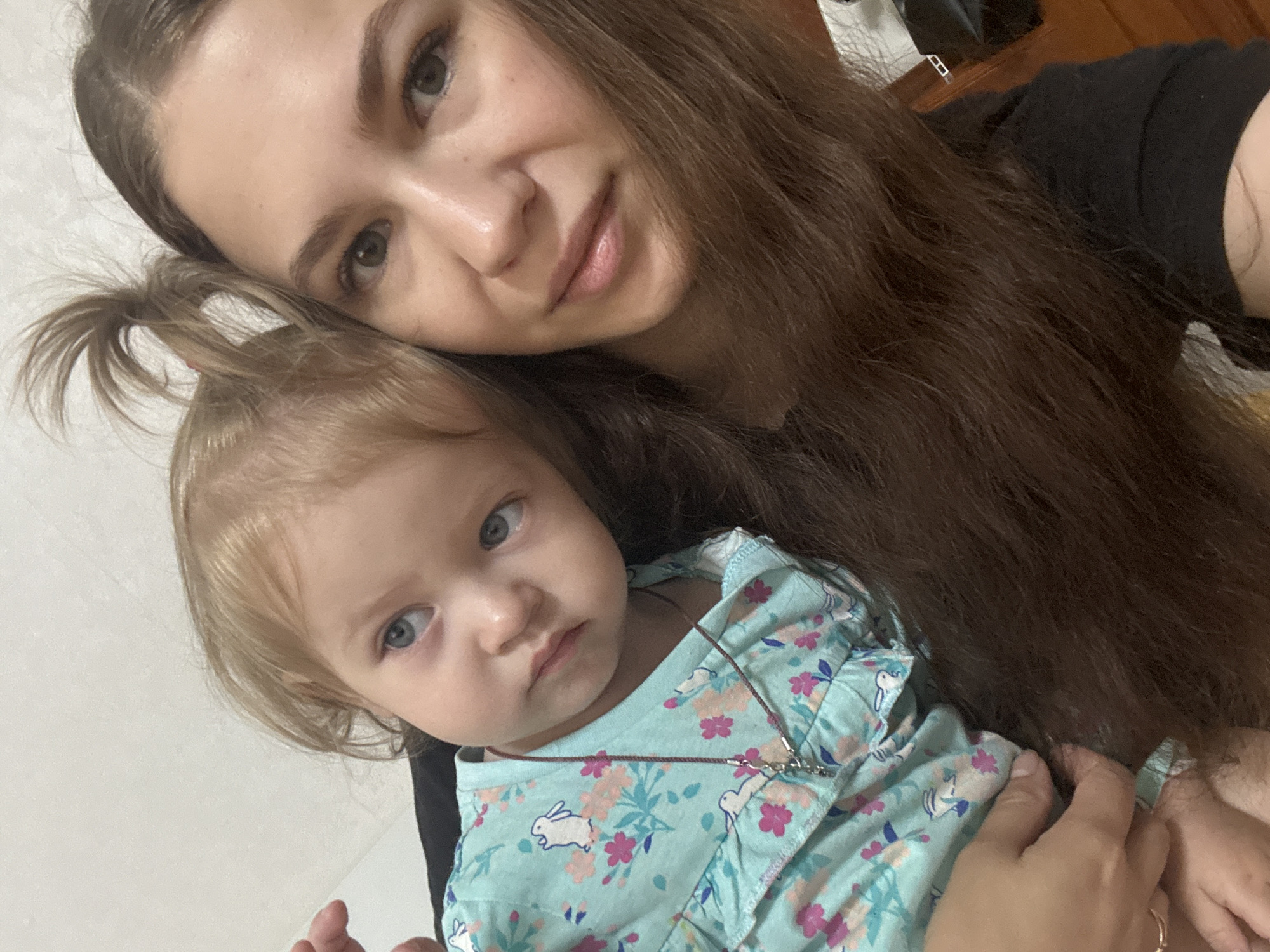 A woman and a young child are in a selfie. The woman has long, dark brown hair and is looking at the camera with a neutral expression. She is wearing a black t-shirt. The child, who appears to be a toddler, is held by the woman and is looking away from the camera with a pensive expression. The child has blonde hair styled in a small ponytail and is wearing a light blue dress with a floral pattern and cartoon rabbits. A thin necklace with a pendant is visible on the child's dress. The background is a plain, light-colored wall. The scene is indoors. The location is not identifiable within the city of Chaikovsky, Russia from the visual information provided in the image.